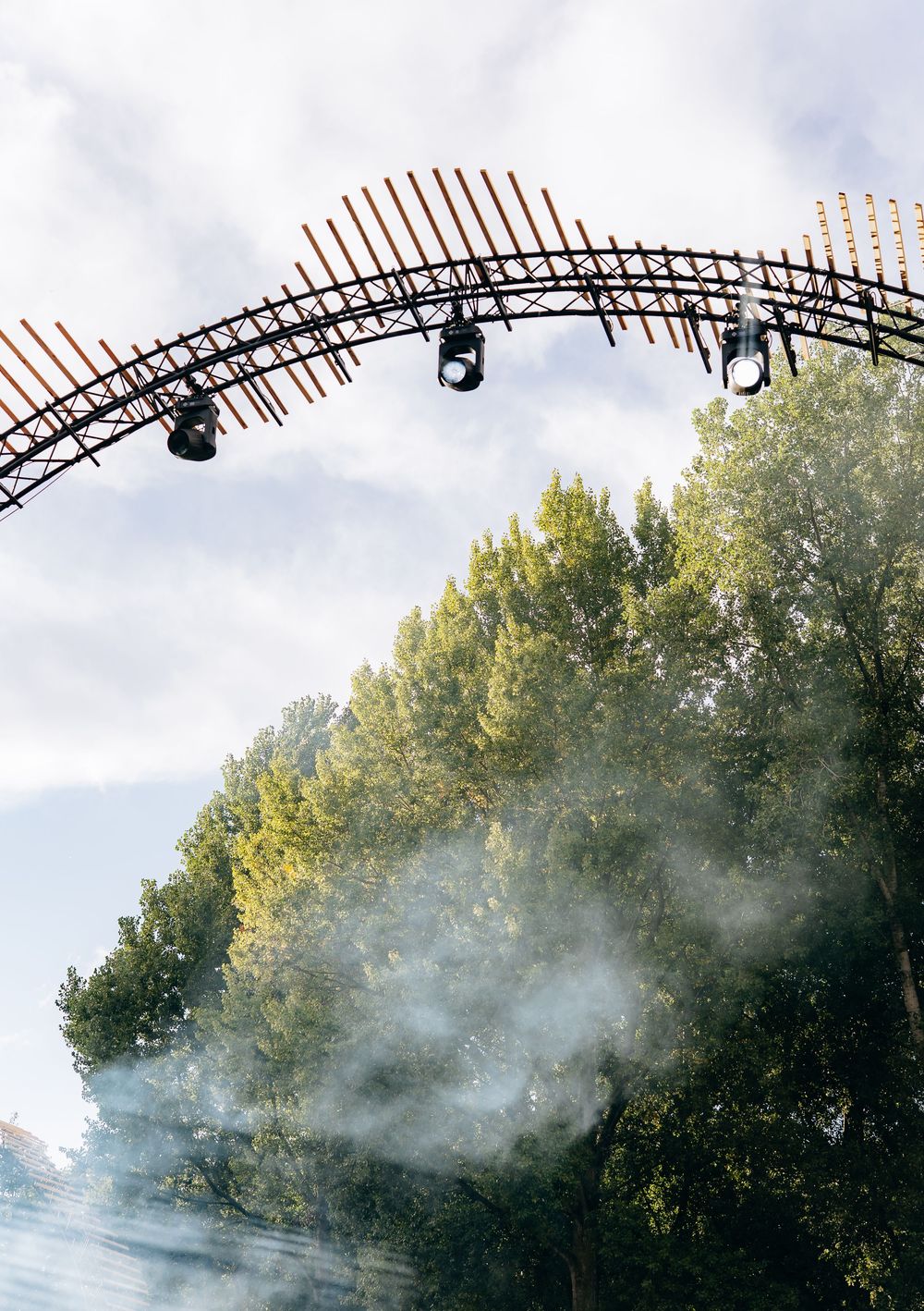 Curved festival stage arch with spotlights over green trees with atmospheric fog below.