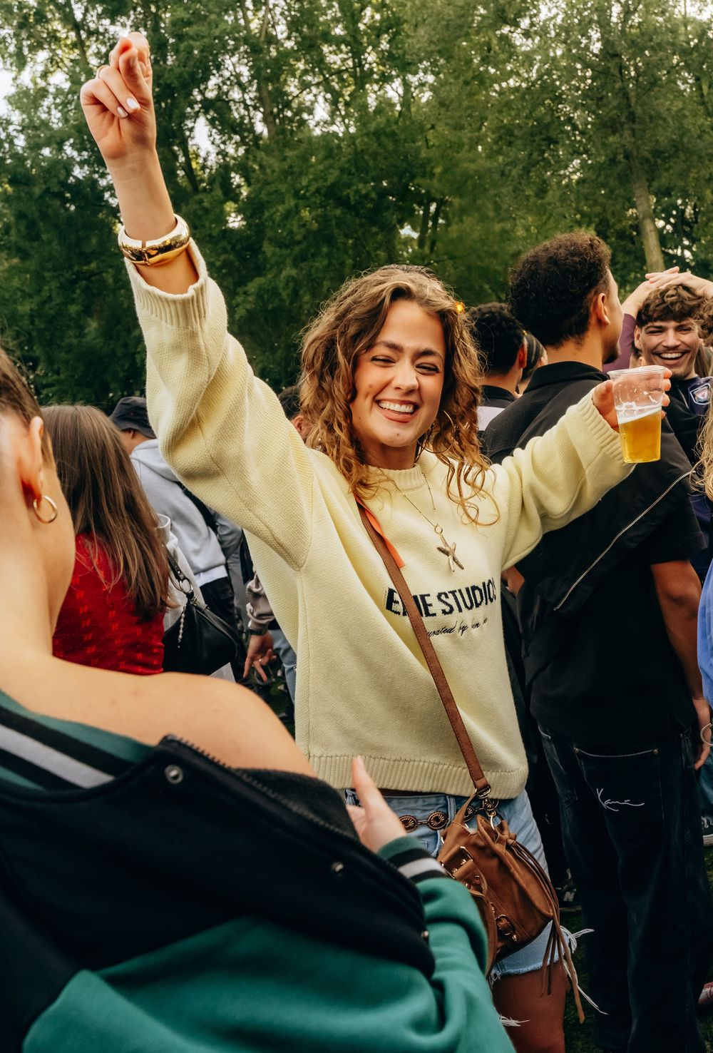 Happy woman dancing with raised arm holding beer at a sunny outdoor music festival.