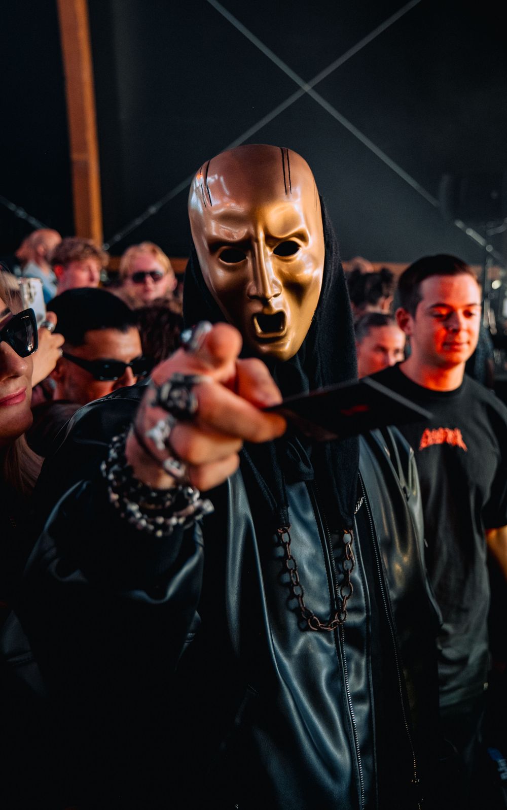 Person wearing a gold theatrical mask and black leather outfit at a dark music event.