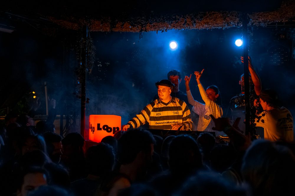 DJ performing at an outdoor night festival stage with blue lights and crowd dancing behind him.