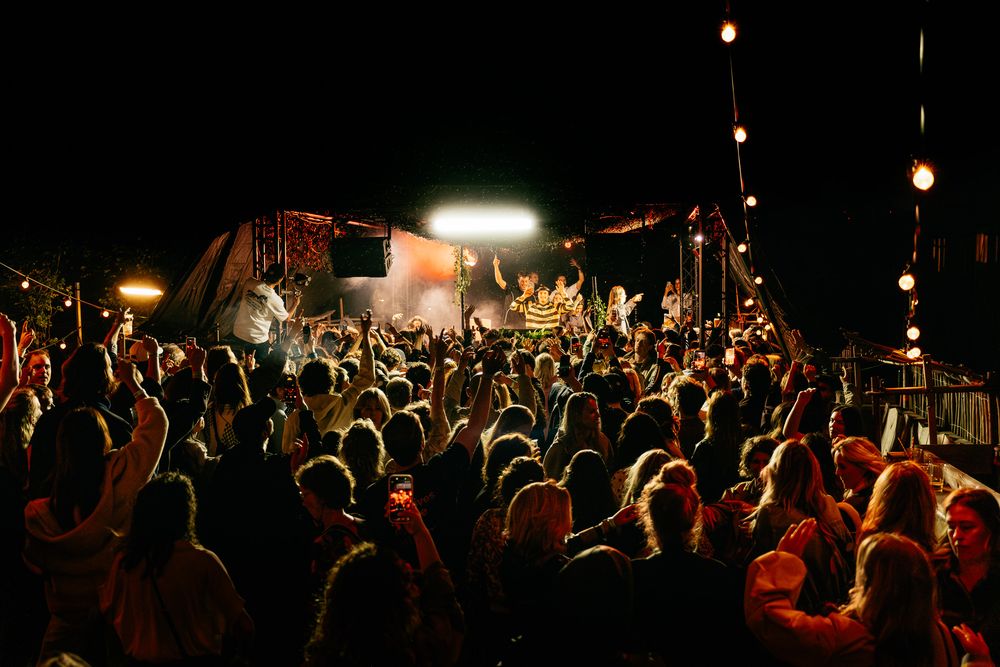 Large crowd gathered at an outdoor night concert with bright stage lights illuminating performers on a decorated stage.