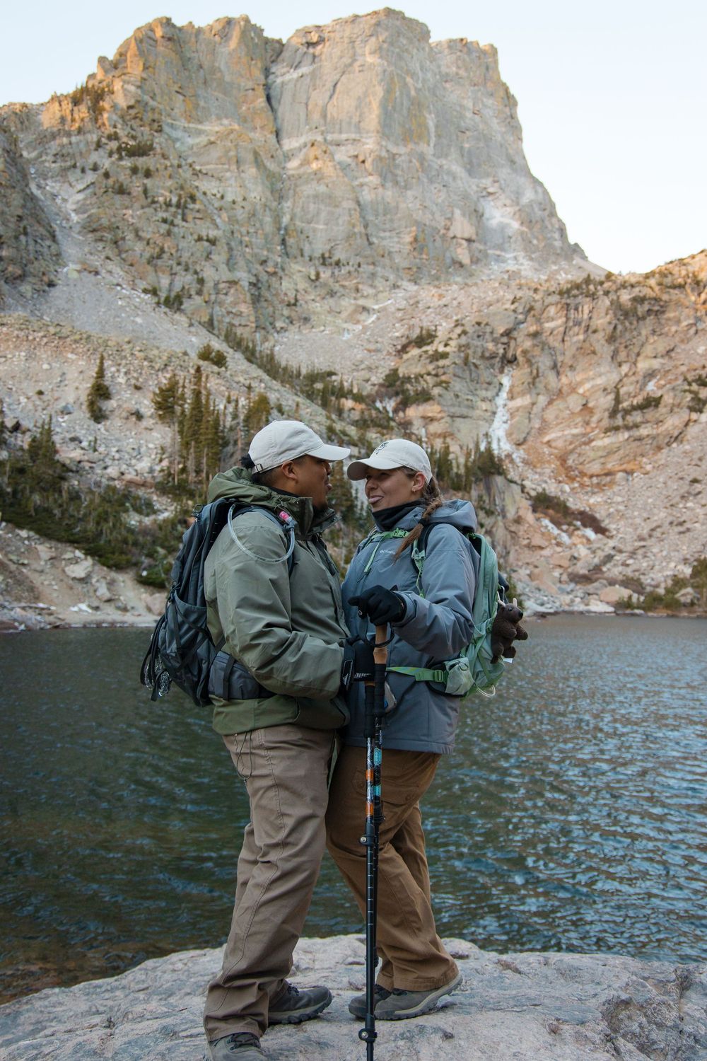 Two hikers with backpacks stand on rocks facing each other near a mountain lake during a trek.