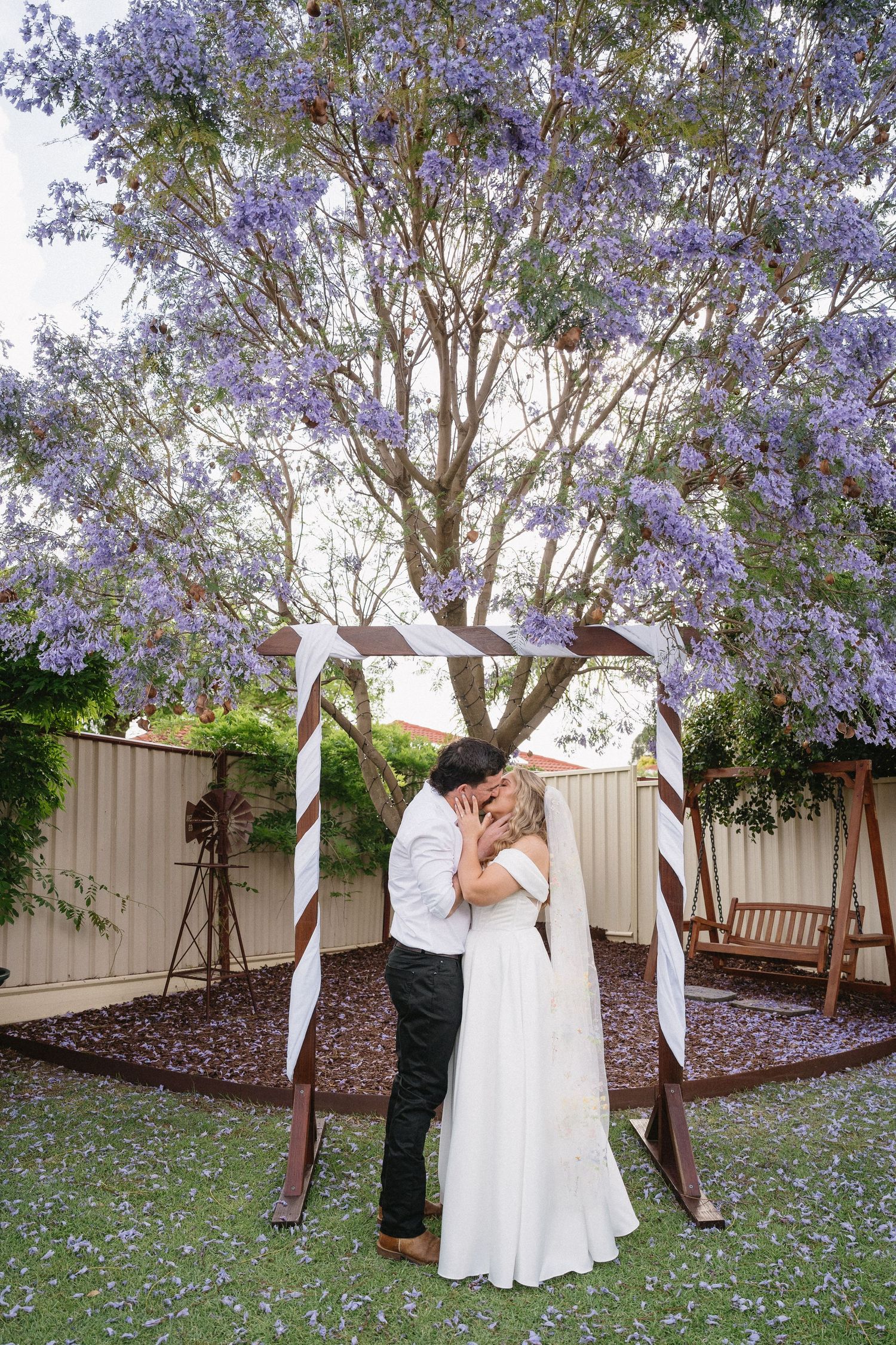 Bride and groom kissing under a wooden arch with a blooming purple jacaranda tree in a garden setting.