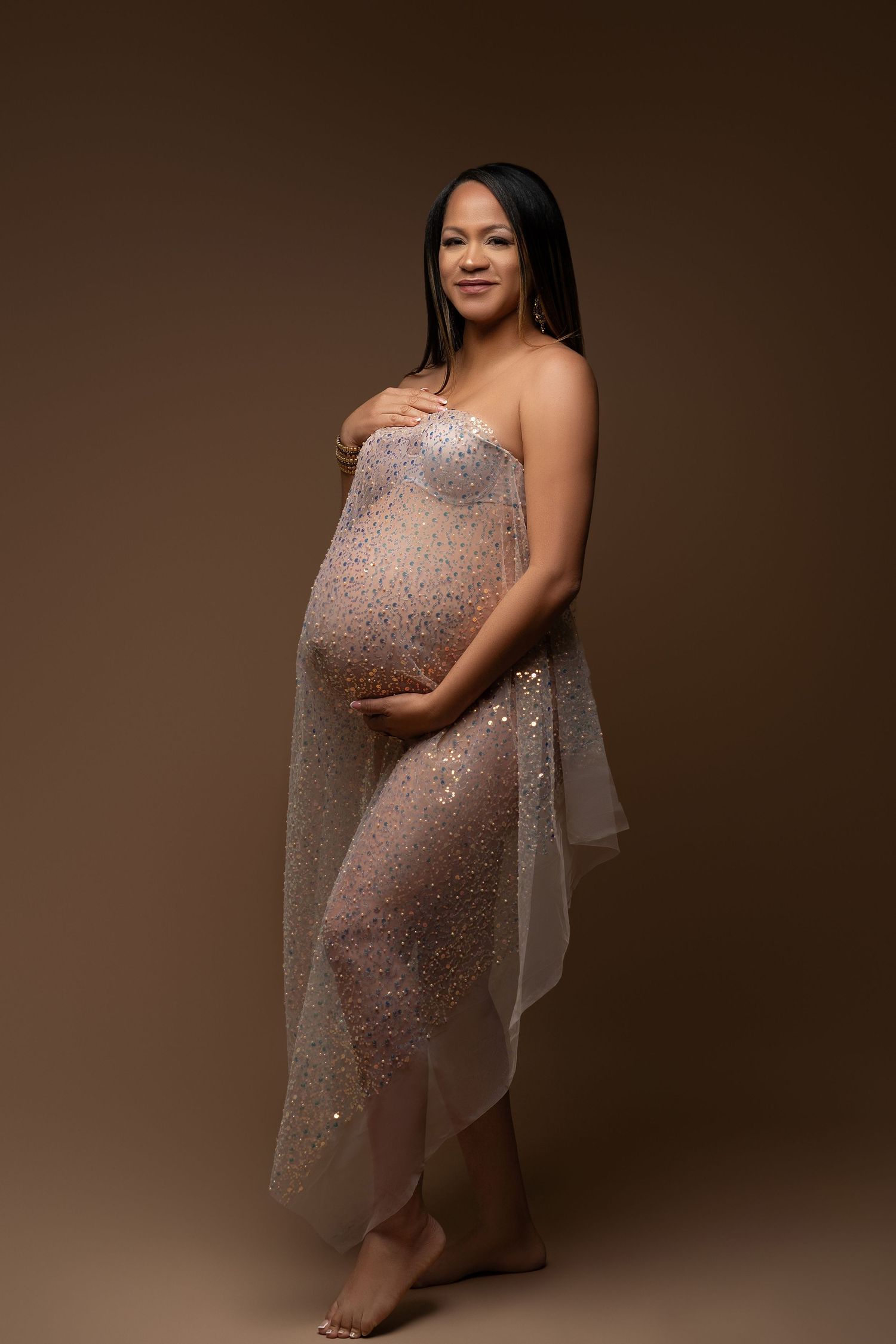 Pregnant woman with curly hair draped in sheer pearl-embellished fabric poses sideways in studio.