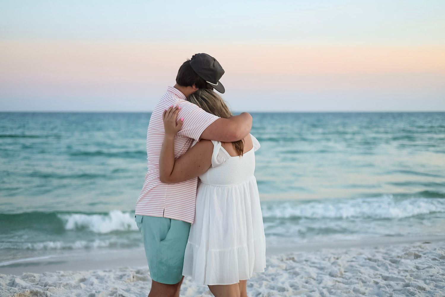 Couple embraces facing the ocean at dusk on a calm sandy beach.