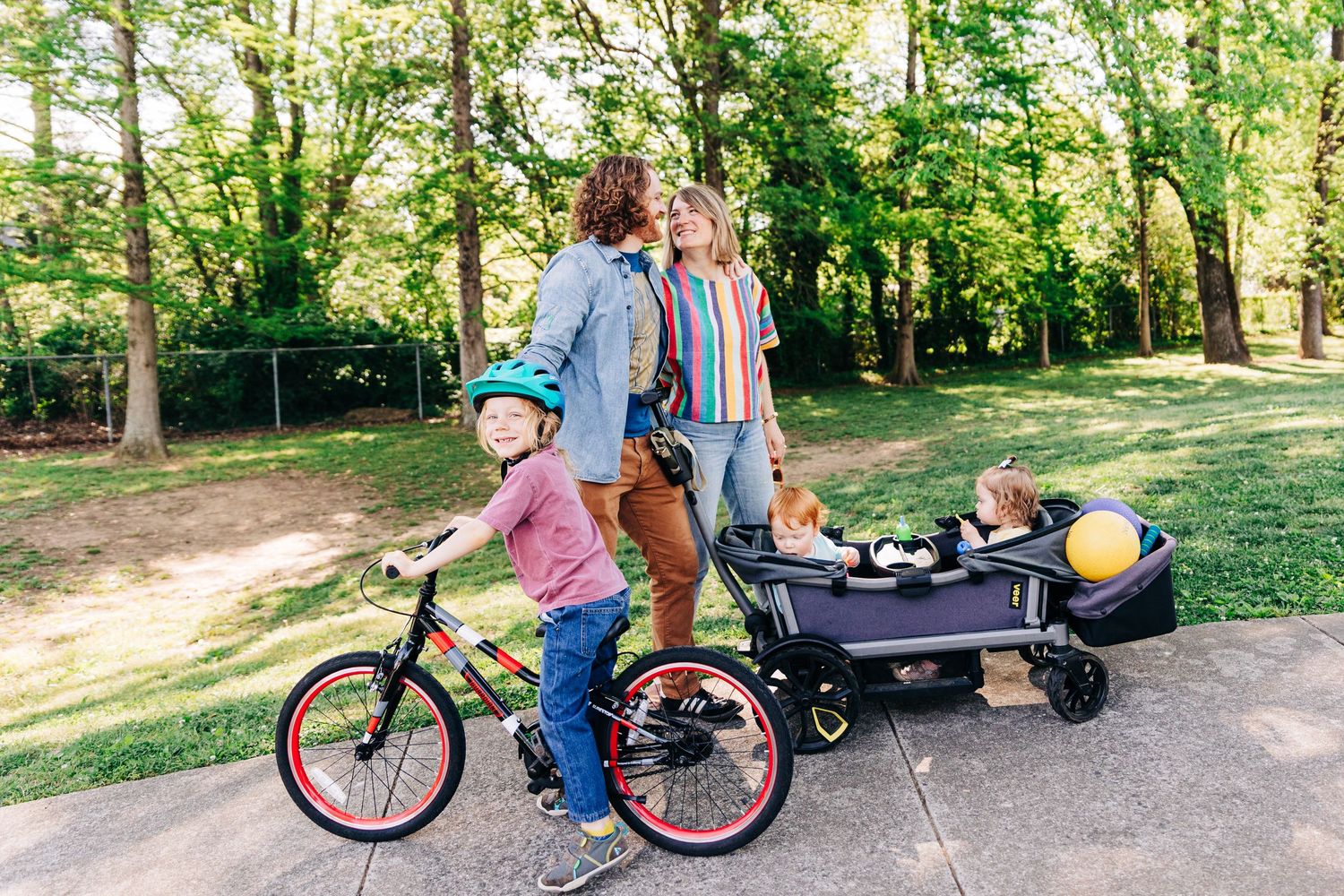 Family enjoying outdoor walk with child on bike and toddlers in stroller wagon on sunny park path.