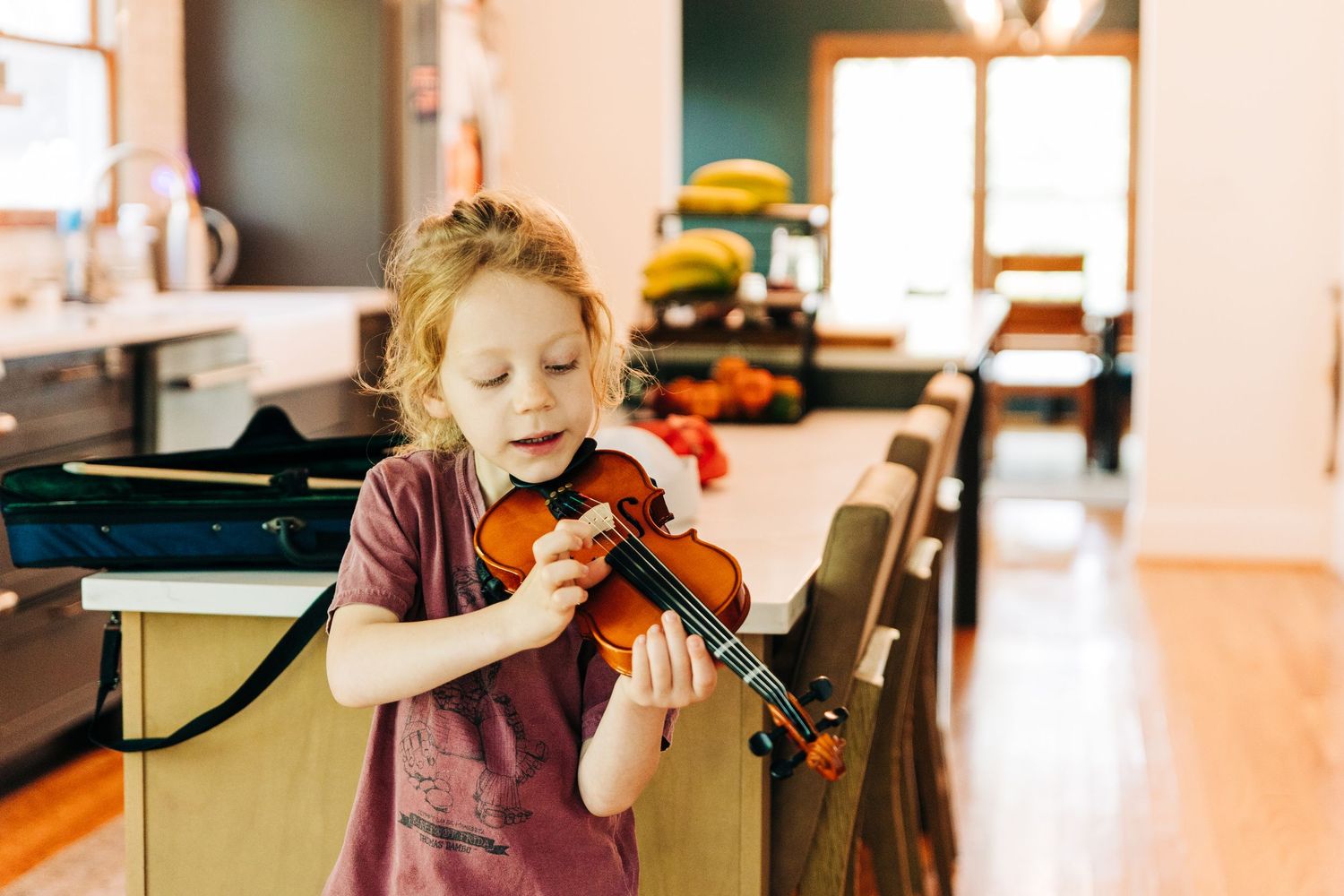 Young child playing a small violin indoors, learning music in a cozy home setting with warm wood floors.