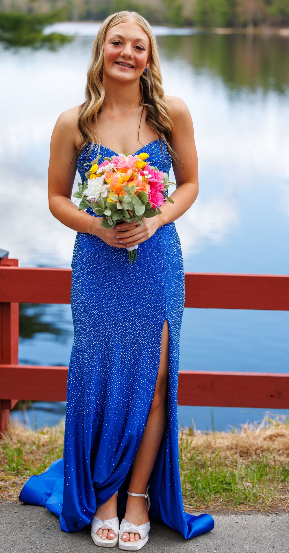 Young woman in royal blue sequined gown holding colorful bouquet, posing by lakeside red fence.
