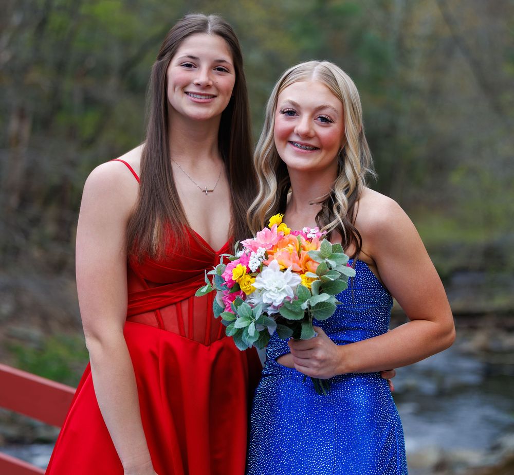 Two young women in formal dresses, one red and one blue, posing together outdoors holding a colorful bouquet.