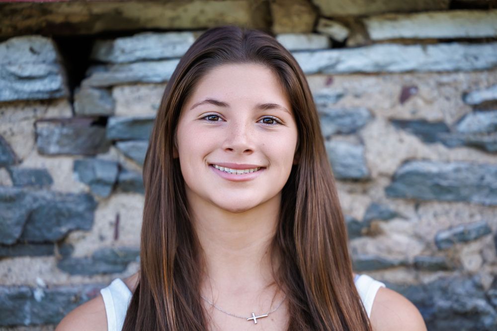 Smiling young woman with brown hair and braces wearing a white top and cross necklace against a stone wall.