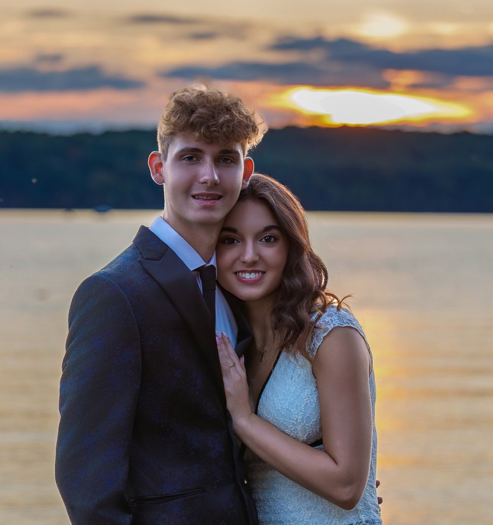 A young couple poses romantically by a lake at sunset, dressed in formal attire for a special occasion.