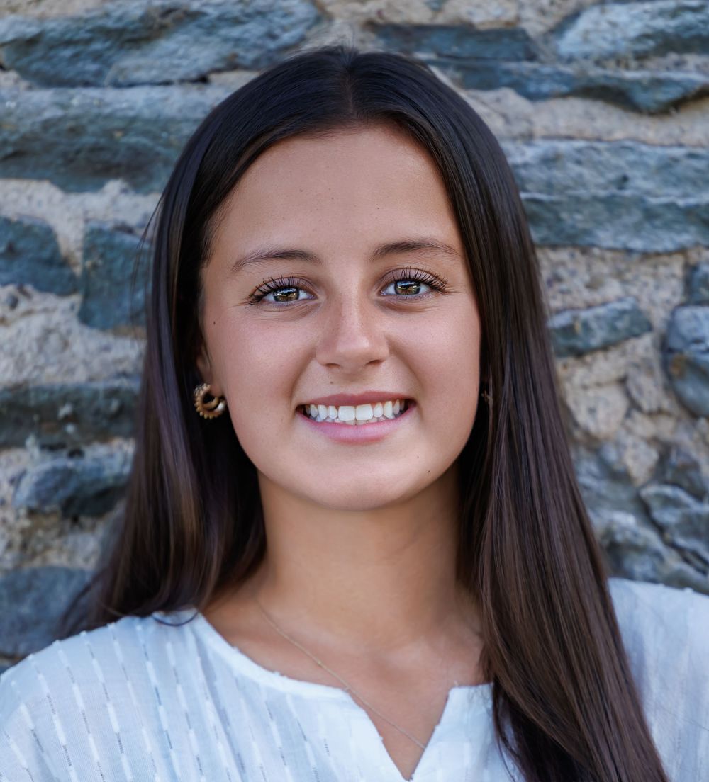 Young woman with long dark hair and blue eyes smiling outdoors against a stone wall background.