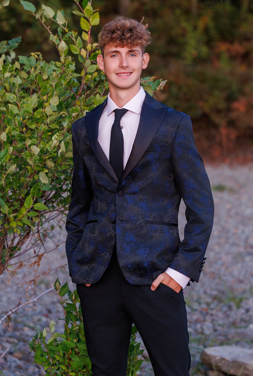 Young man in navy blue floral tuxedo jacket with black lapels and tie, posing outdoors near green foliage.