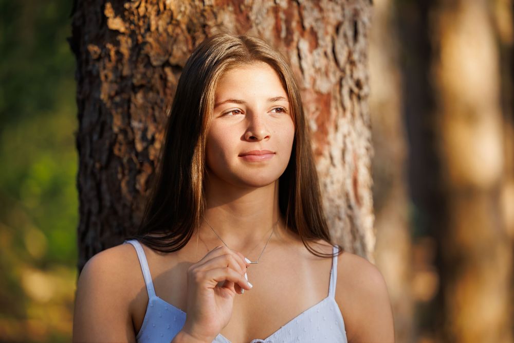 Young woman with long brown hair leaning against a tree trunk in golden sunlight outdoors.