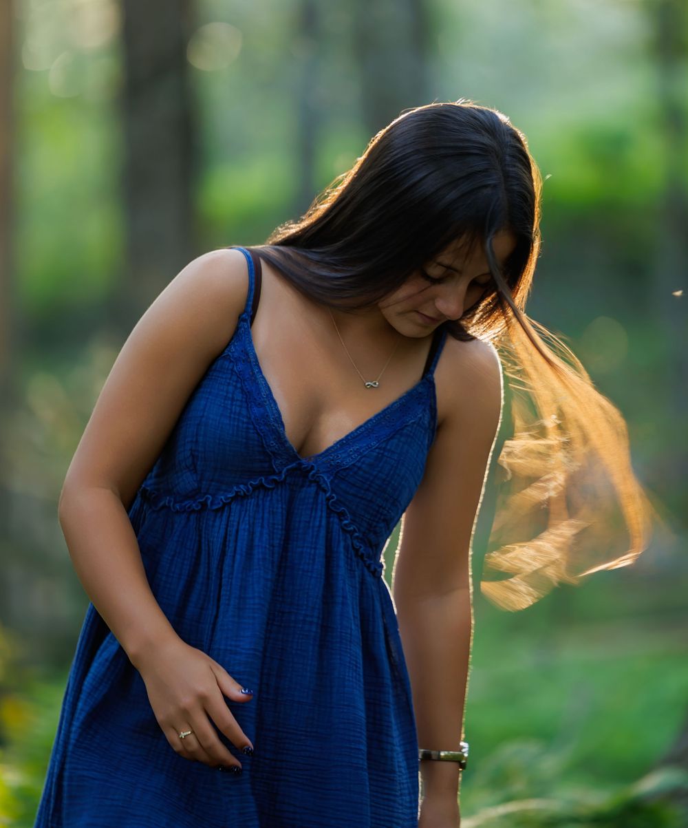 Young woman in blue sundress standing in sunlit forest with hair blowing in breeze.
