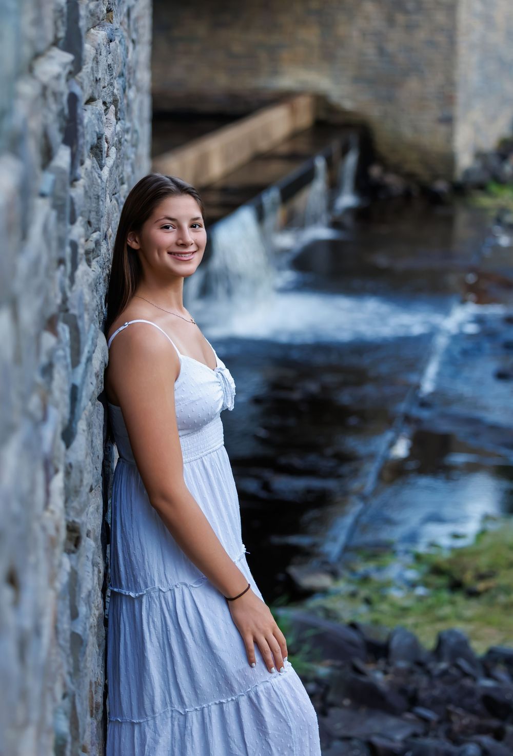 Young woman in white sundress smiling by stone wall with waterfall backdrop.