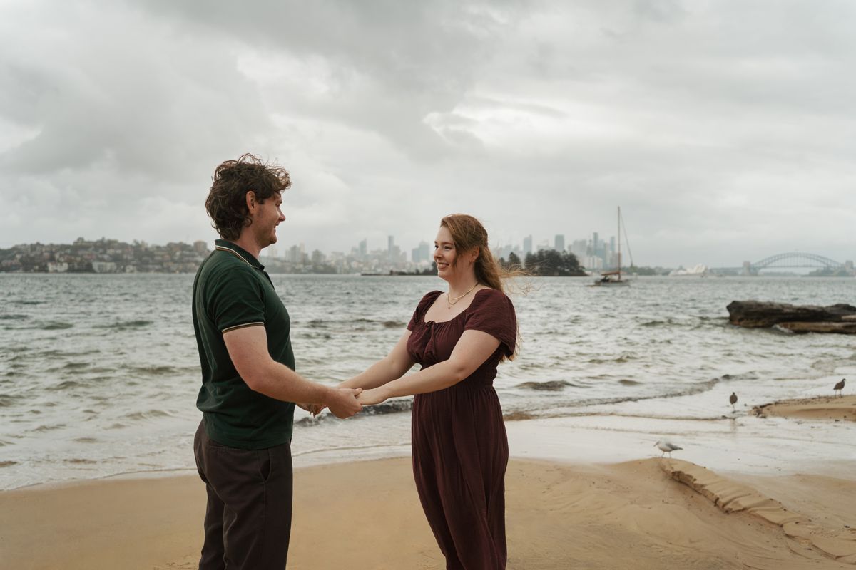 Couple holding hands on Sydney beach with harbour bridge and city skyline in background on cloudy day proposal photoshoot