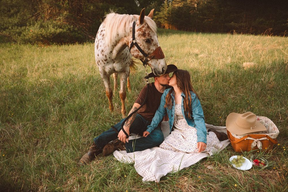 Romantic couple sitting together on a blanket in a field during a picnic with a horse standing nearby.