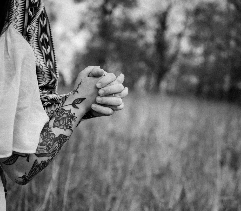 Black and white photo of clasped hands with floral tattoo sleeve outdoors in a field.