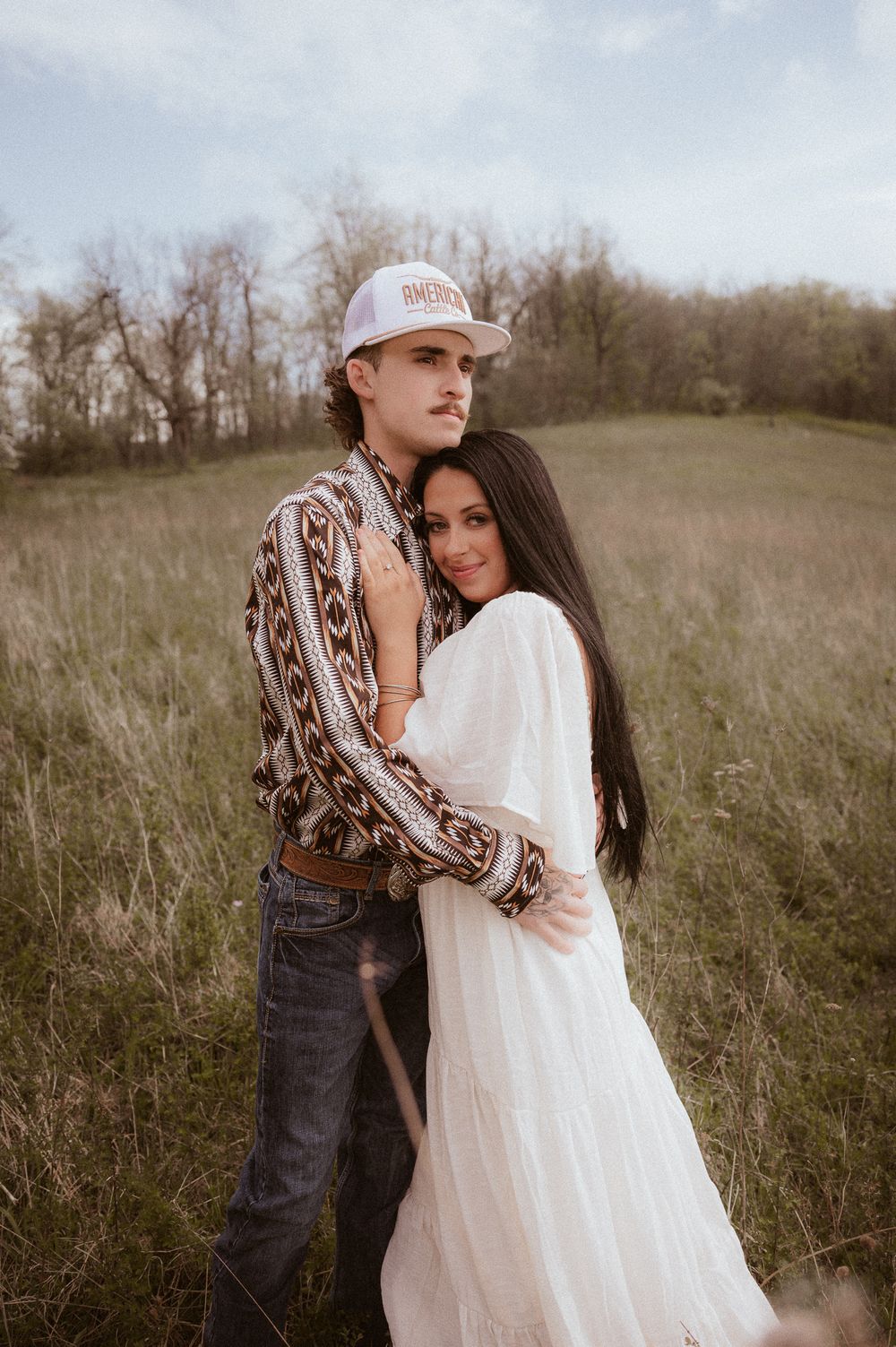 A couple embraces in a grassy field, woman in white dress, man in patterned shirt and trucker hat.