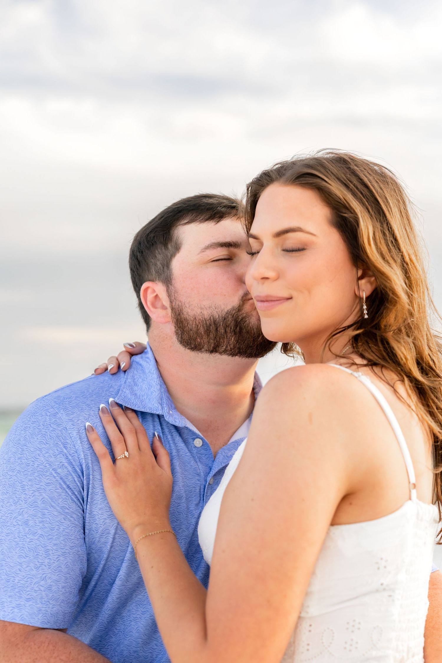 A man in a blue polo kisses a smiling woman in a white dress on the cheek at a beach.