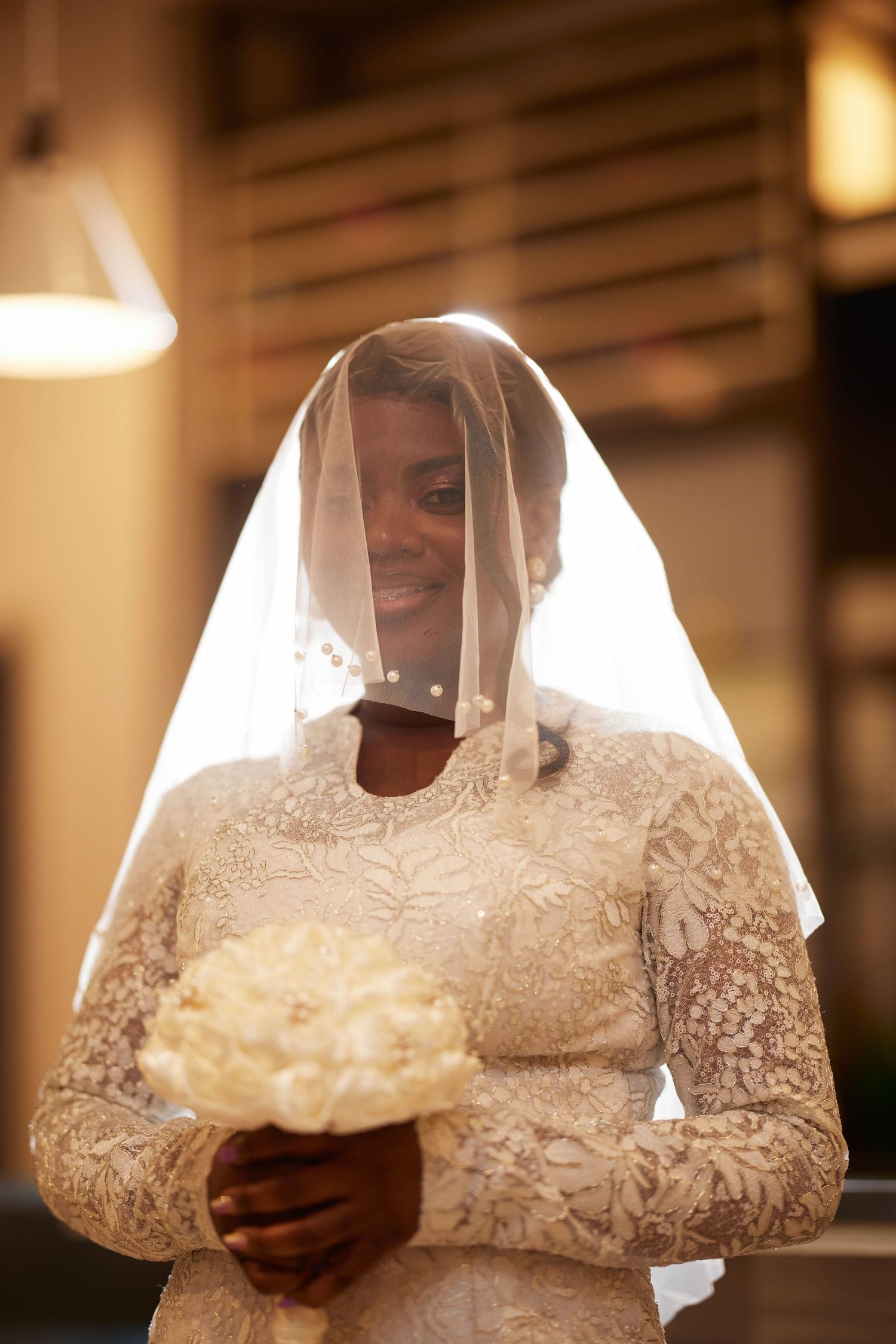 Smiling Black bride in lace wedding dress and veil holds white bouquet under warm light.