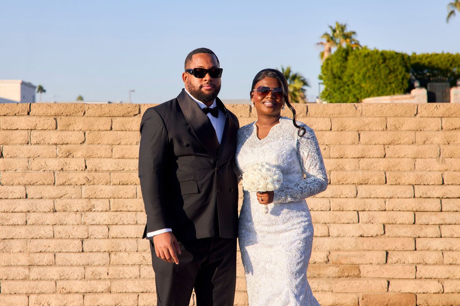 Bride and groom posing in front of a brick wall, bride in white lace gown holding bouquet, groom in black suit.