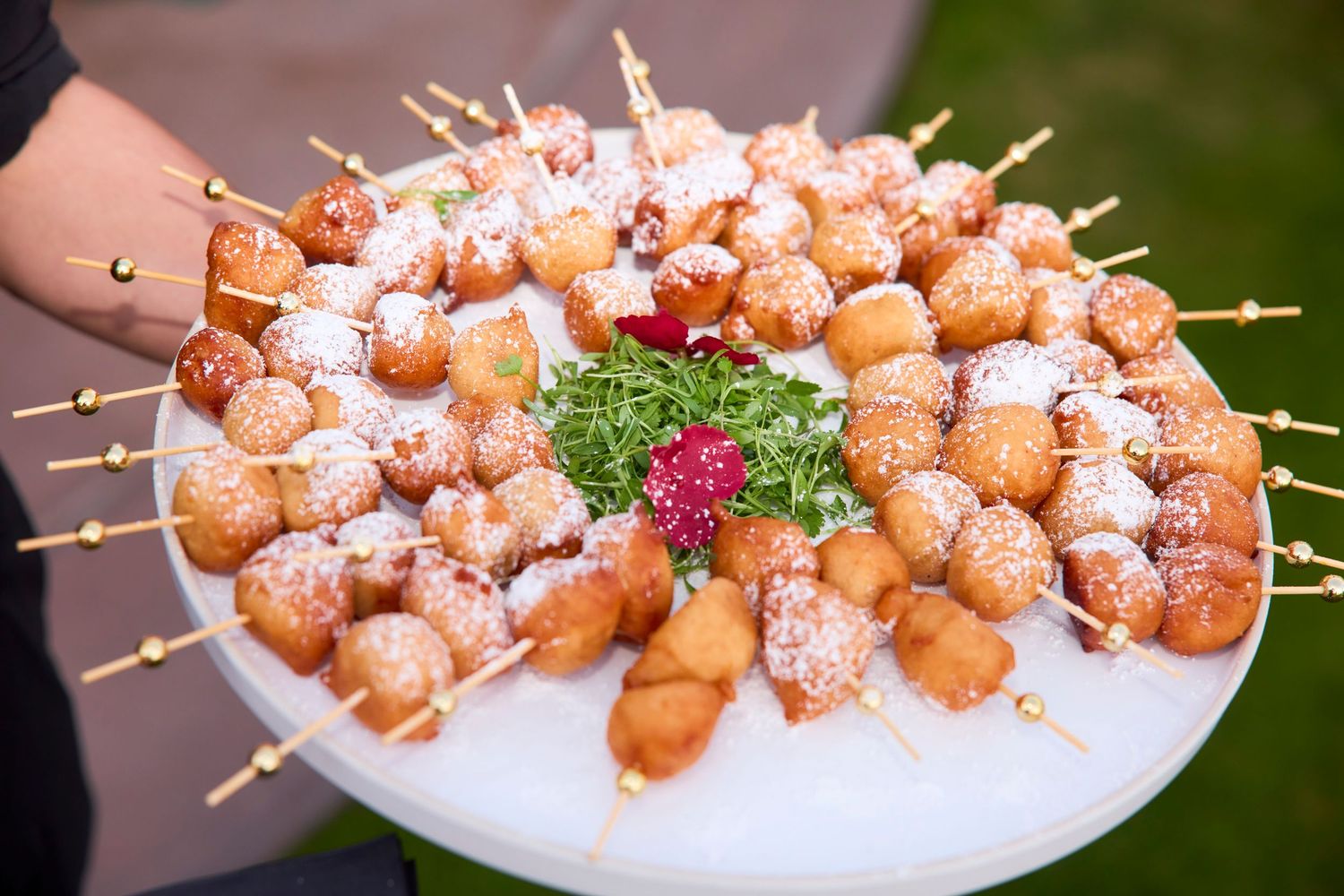 Platter of powdered sugar donut holes on skewers garnished with fresh greens and berries.