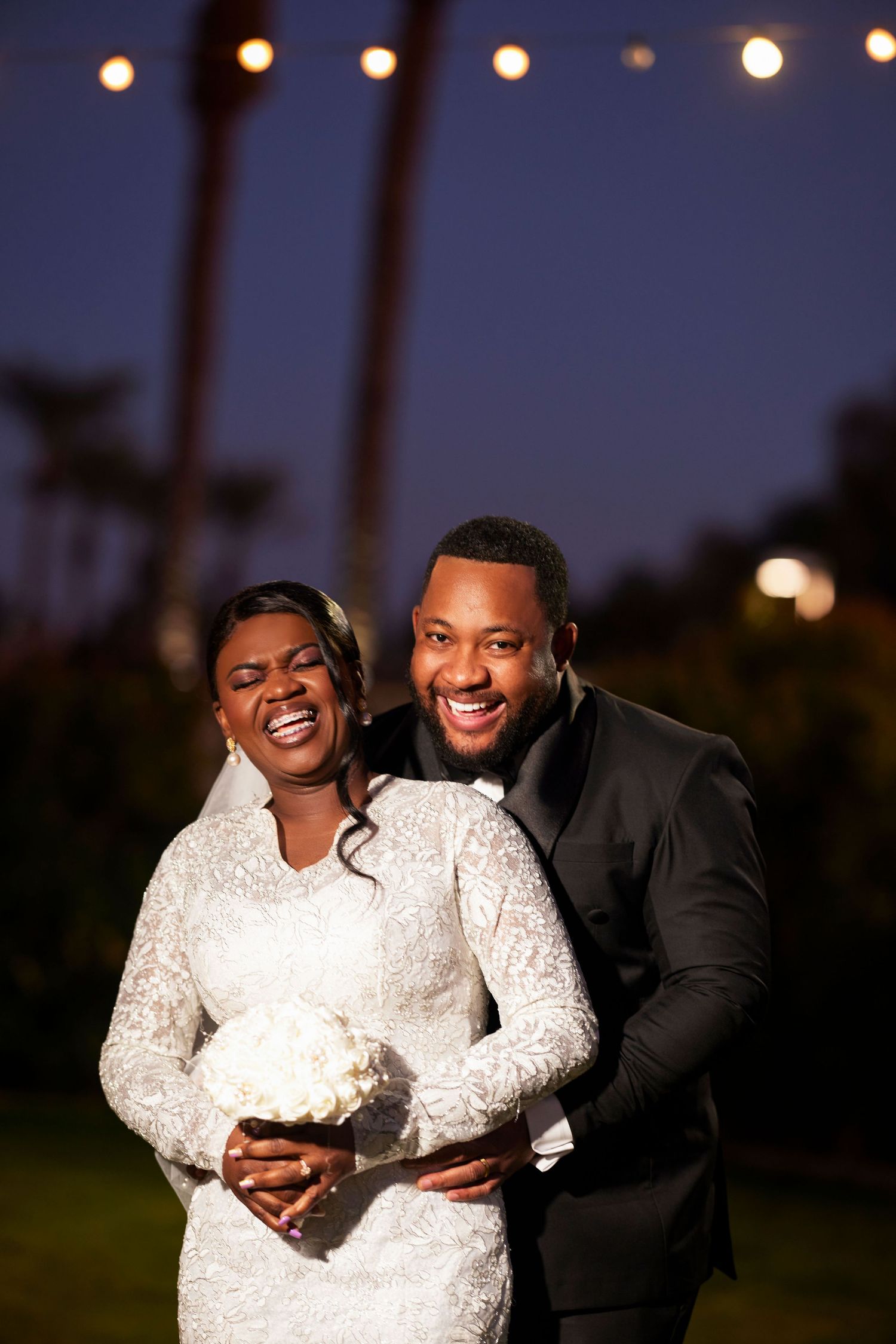 Happy newlywed couple in wedding attire smiling outdoors at night under string lights, bride holding white bouquet.