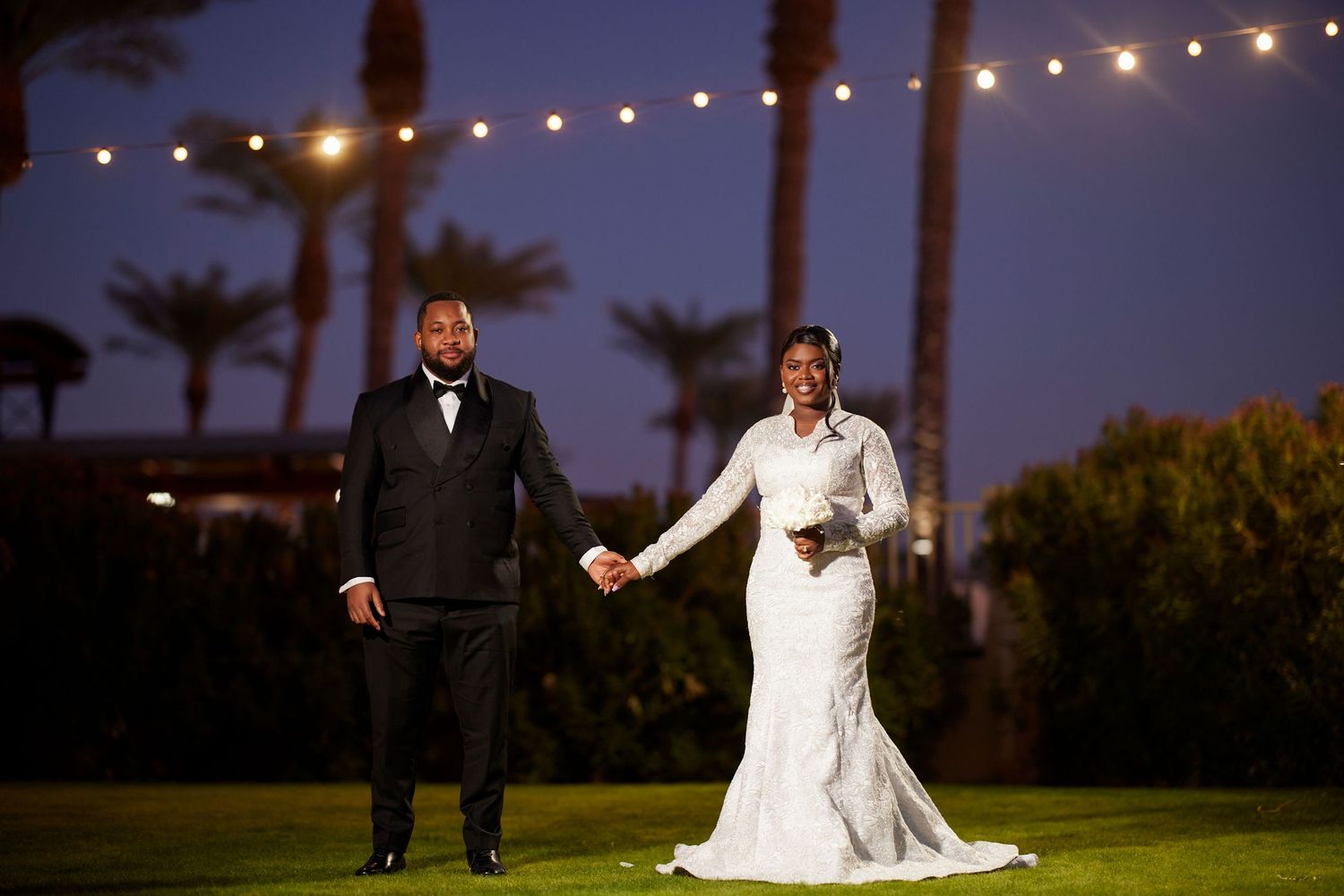 Elegant bride and groom holding hands on a lawn at dusk, with palm trees and string lights glowing behind them.