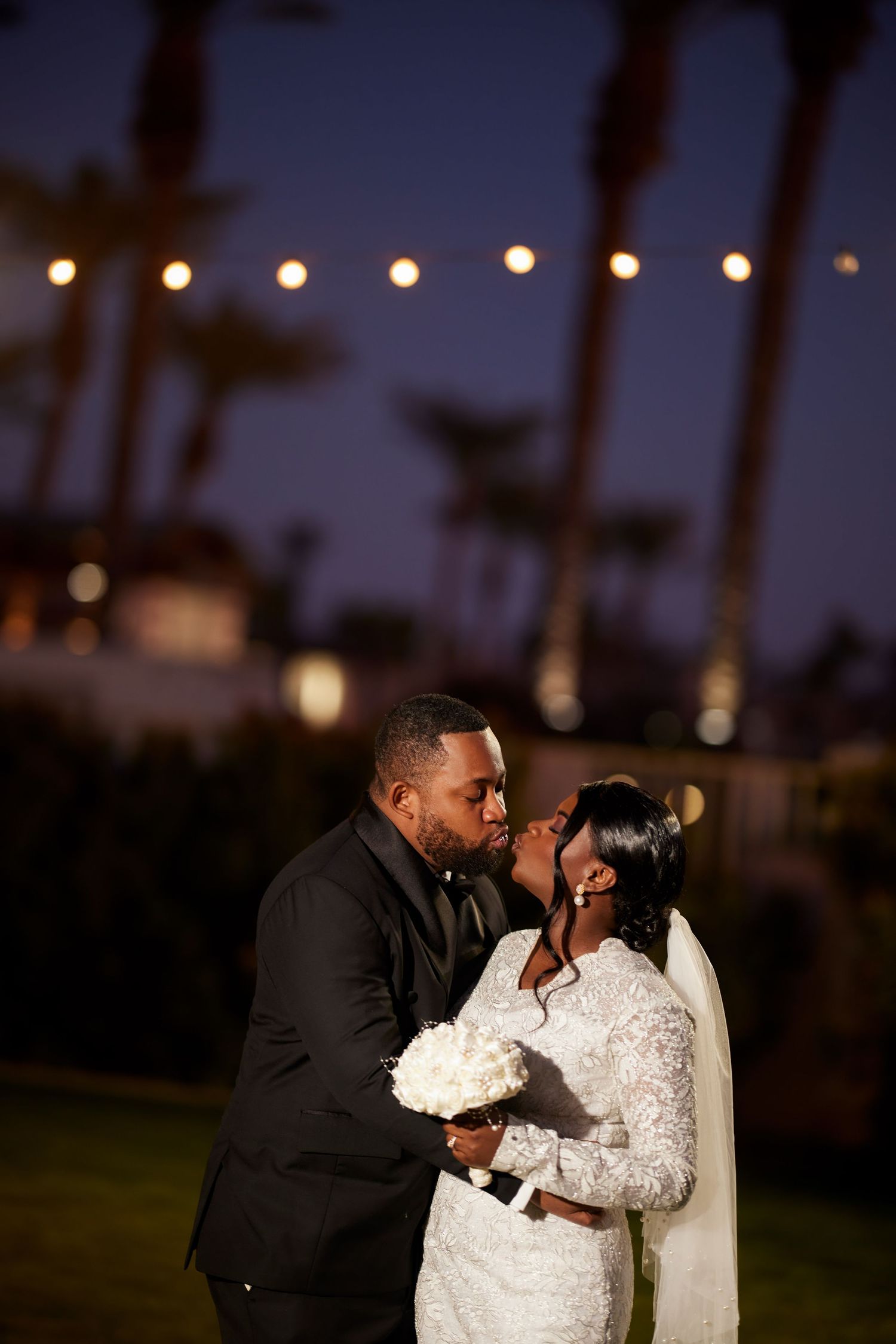 A bride and groom share a romantic kiss at night, with string lights and palm trees glowing softly in the background.
