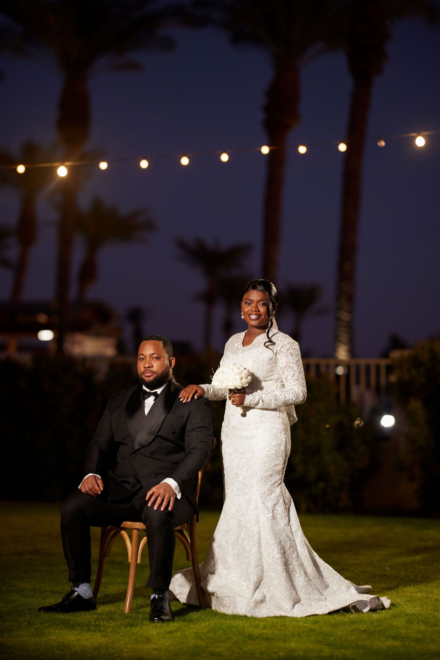 Elegant bride and groom posing outdoors at night, with string lights and palm trees in the background.
