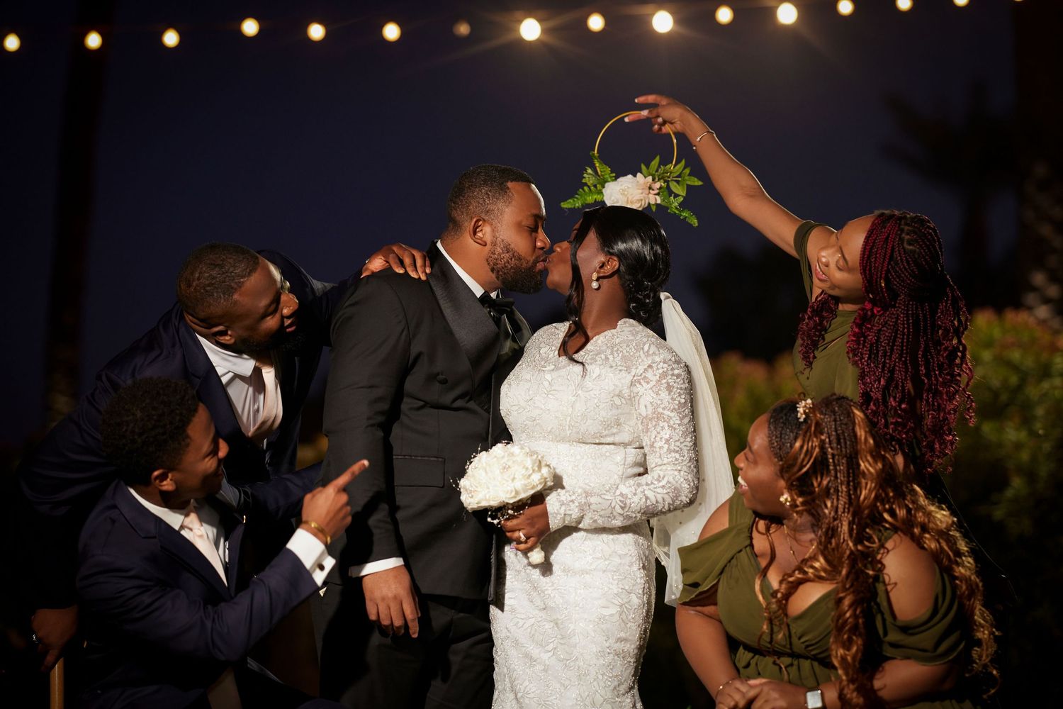 A joyful wedding couple shares a kiss surrounded by celebrating friends under string lights at an outdoor evening ceremony.