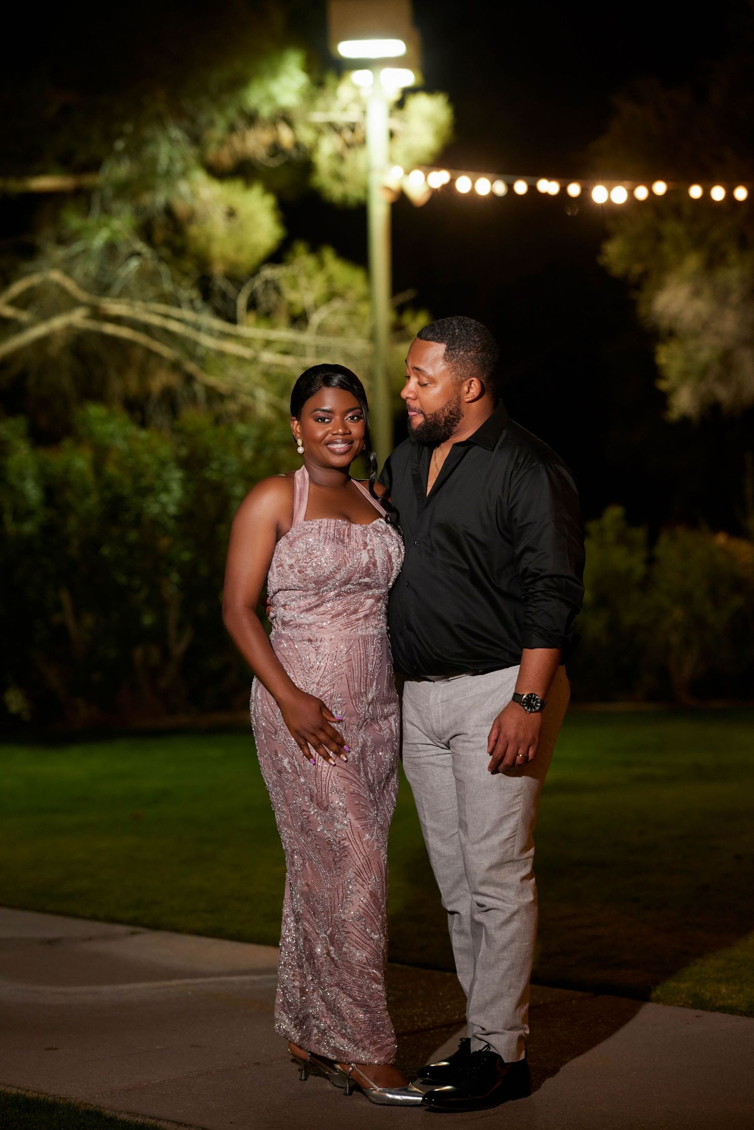 Elegant couple posing outdoors at night, woman in pink lace gown and man in black shirt with gray pants.