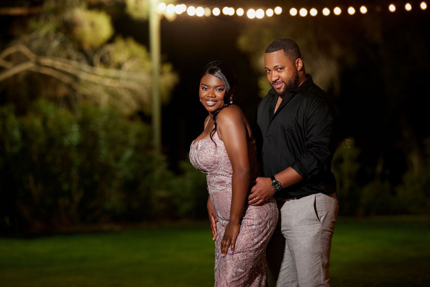 Elegant couple posing outdoors at night, woman in pink gown and man in black shirt, with string lights in background.