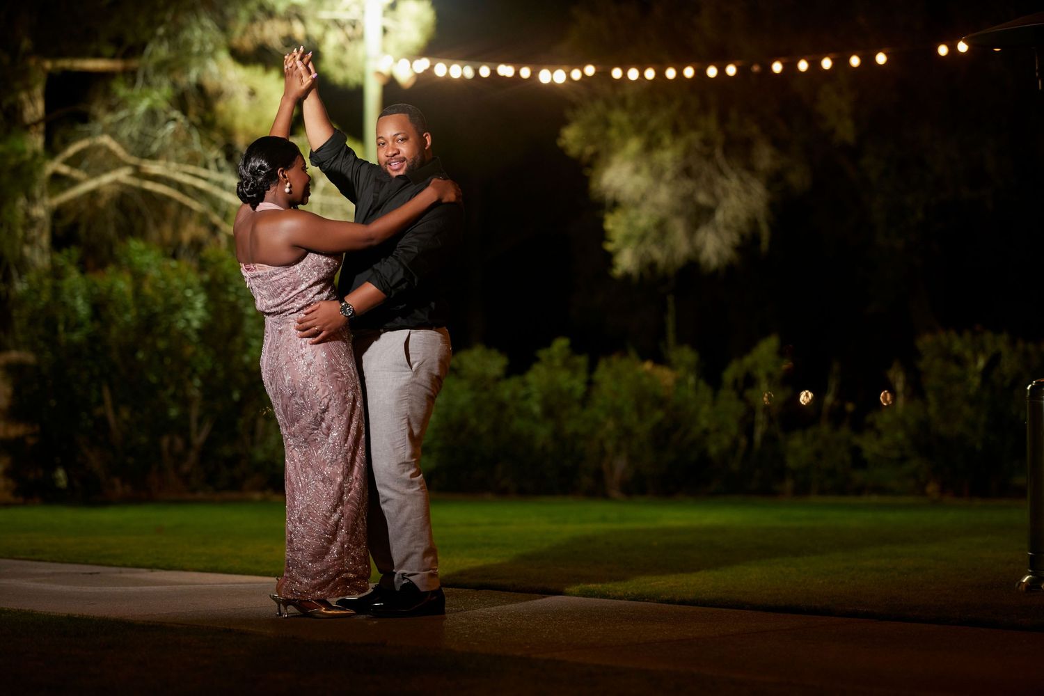 Couple dancing romantically outdoors at night under string lights surrounded by lush greenery.