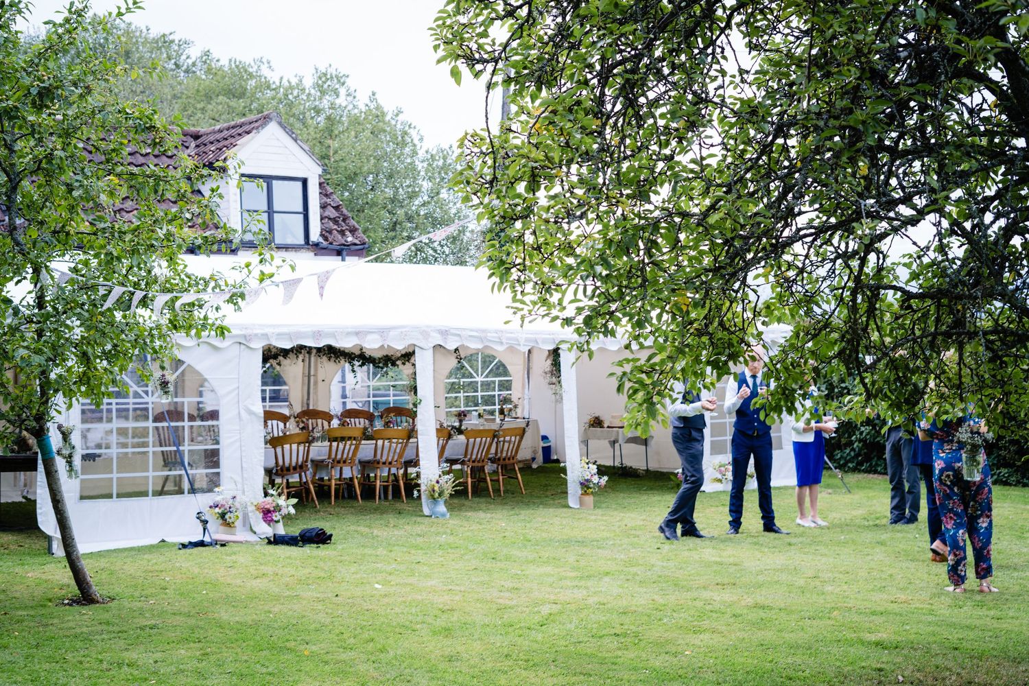 Outdoor wedding reception with a white marquee tent, wooden chairs, and guests socializing on a green lawn.