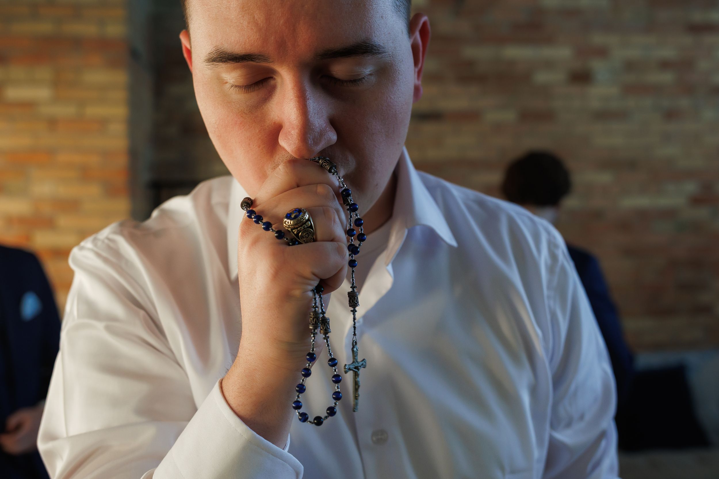 A man in a white dress shirt prays with rosary beads at a wedding ceremony.