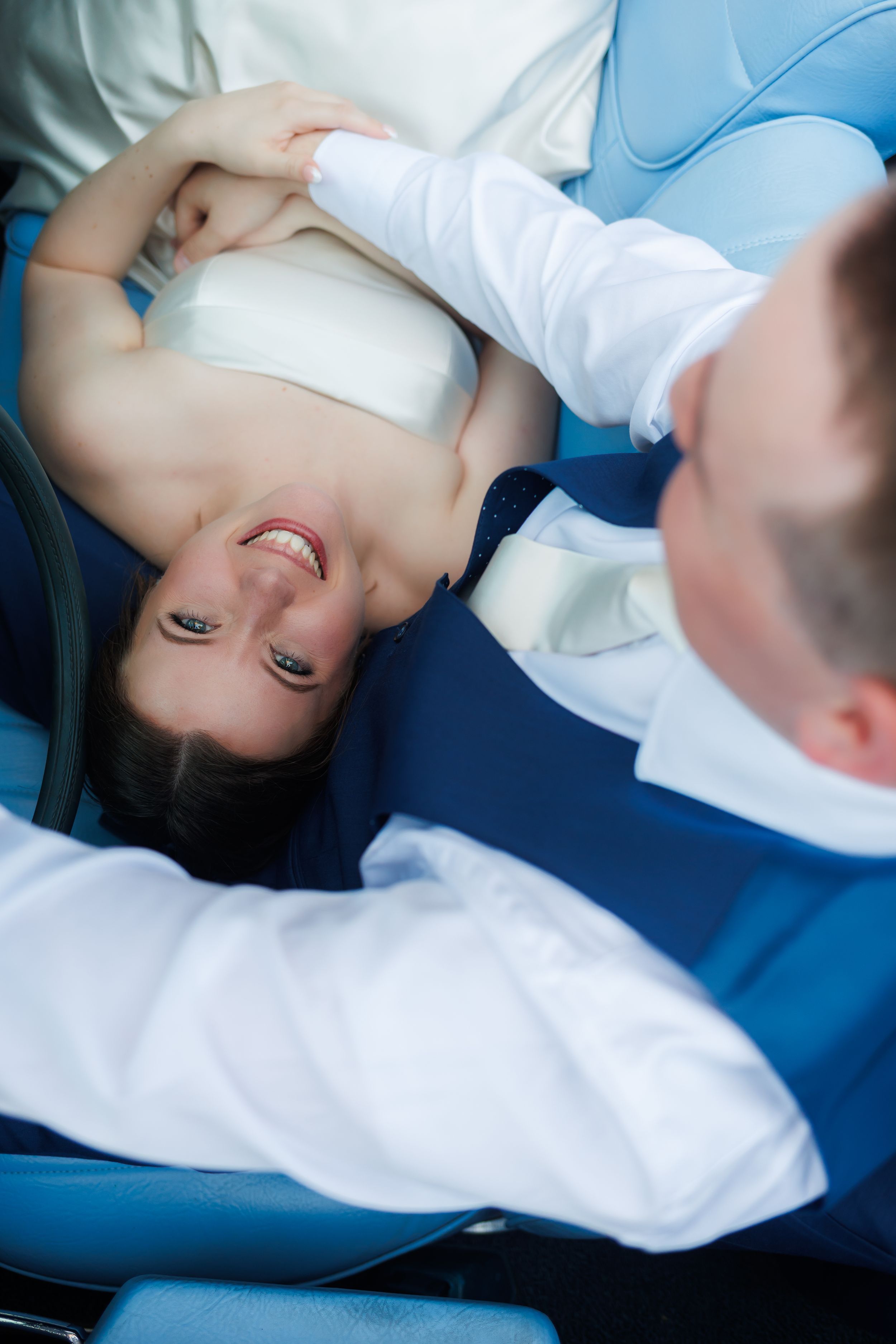 A smiling bride reclines in a dental chair while a dentist in a blue vest examines her teeth before her wedding day.