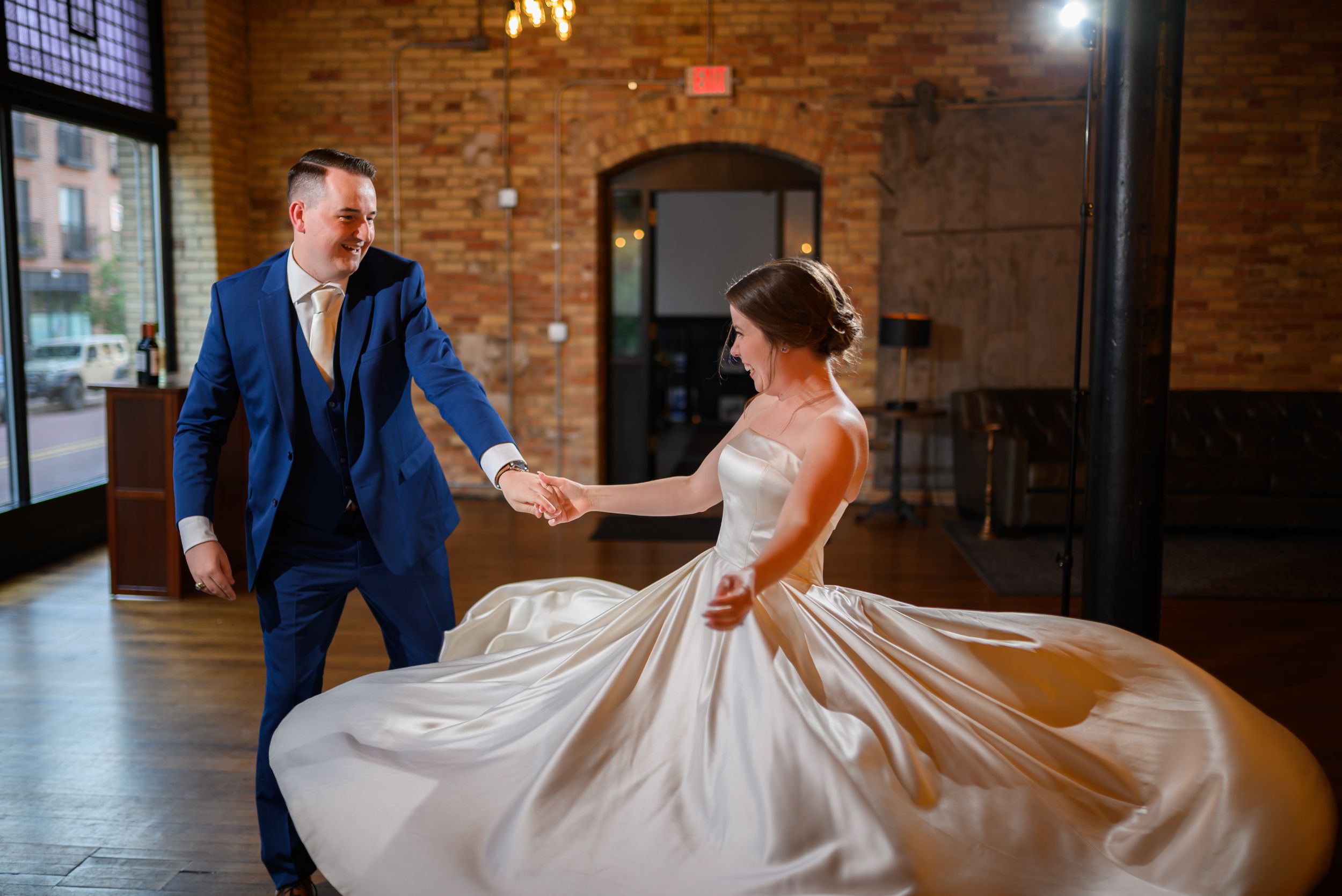 Bride and groom share a joyful first dance, spinning on the dance floor of a rustic brick-walled wedding venue.