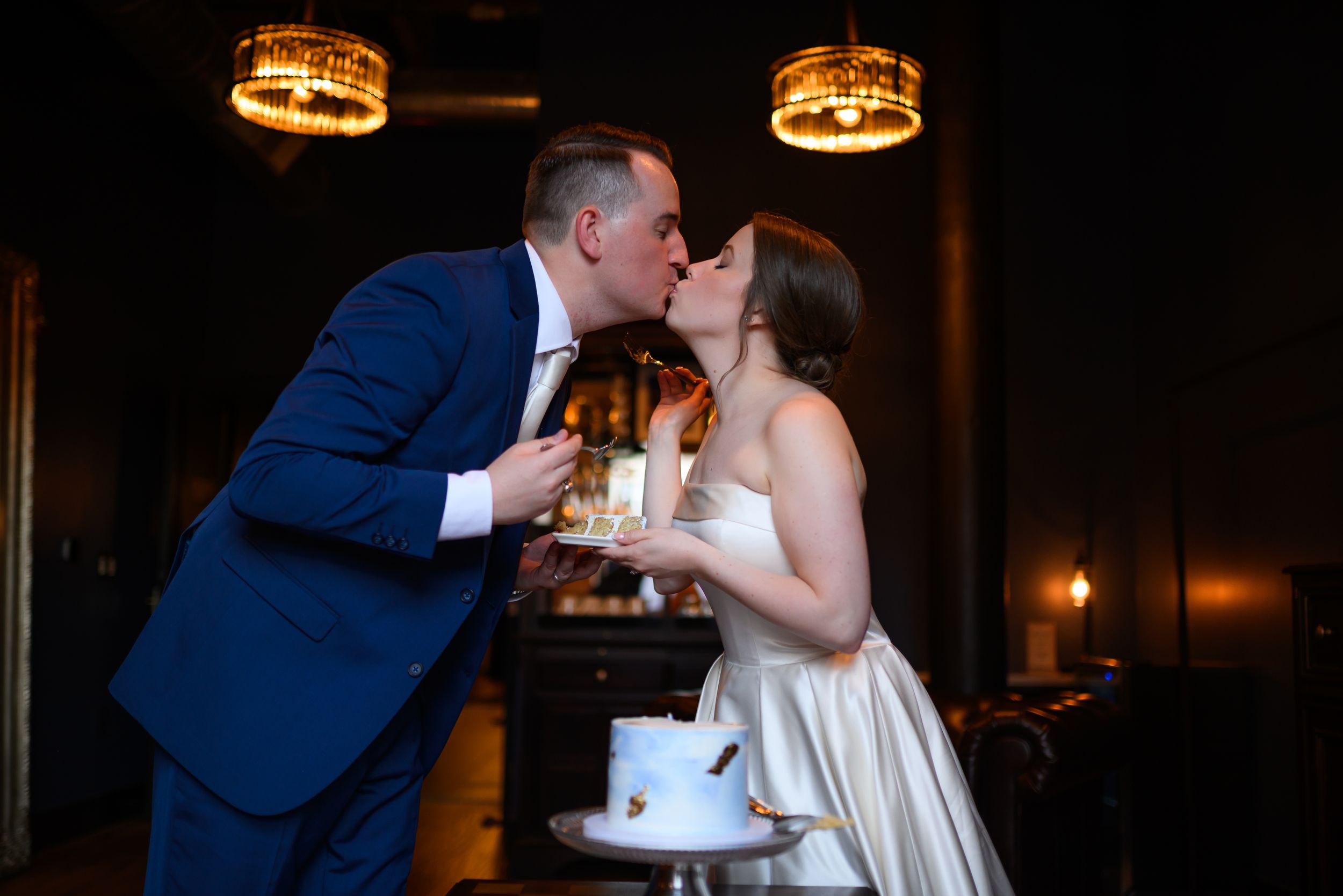 Newlyweds share a romantic kiss at their wedding reception, groom in blue suit, bride in white gown with small cake nearby.