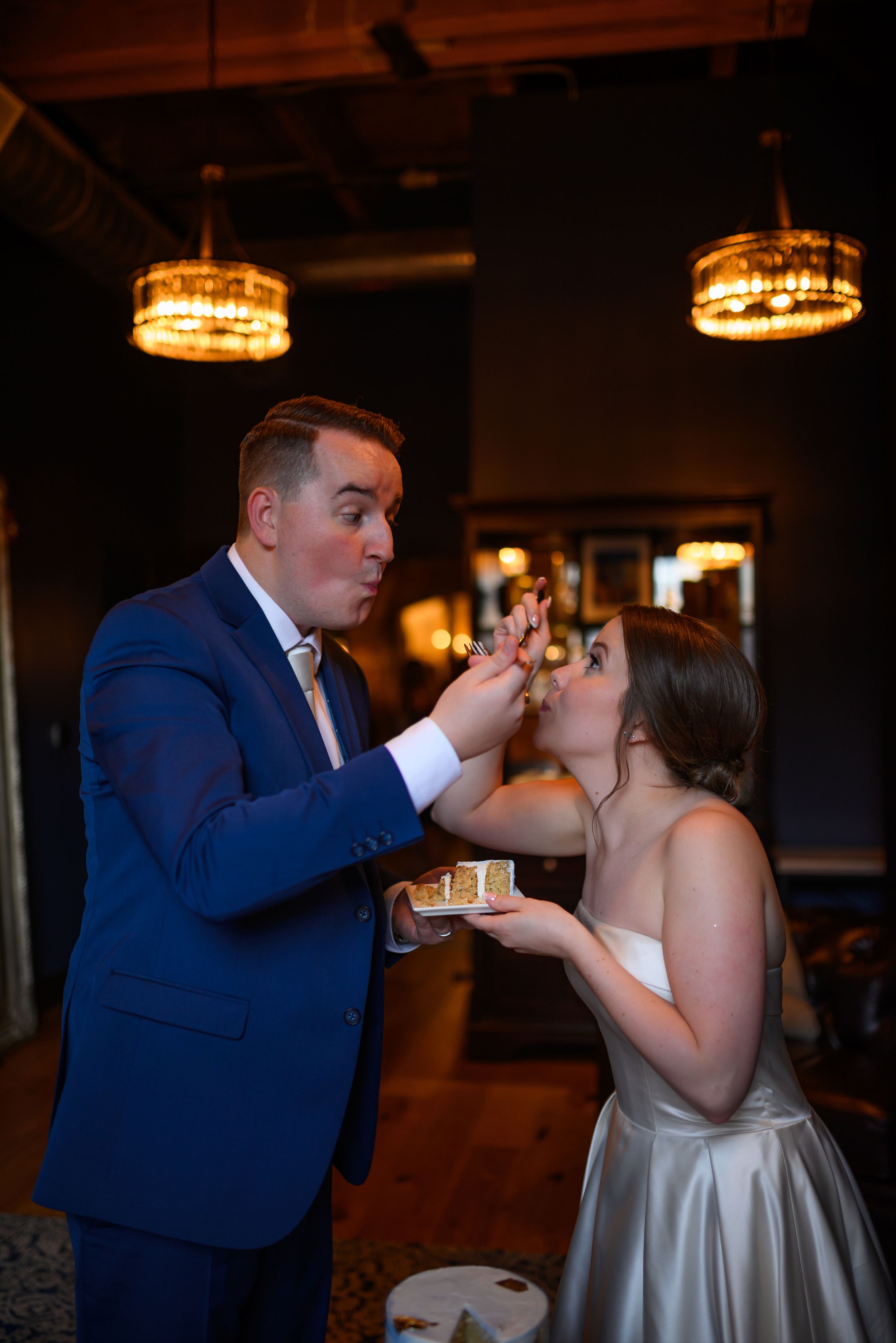 Groom in blue suit feeds wedding cake to bride in strapless gown at elegant reception venue with chandelier lighting.