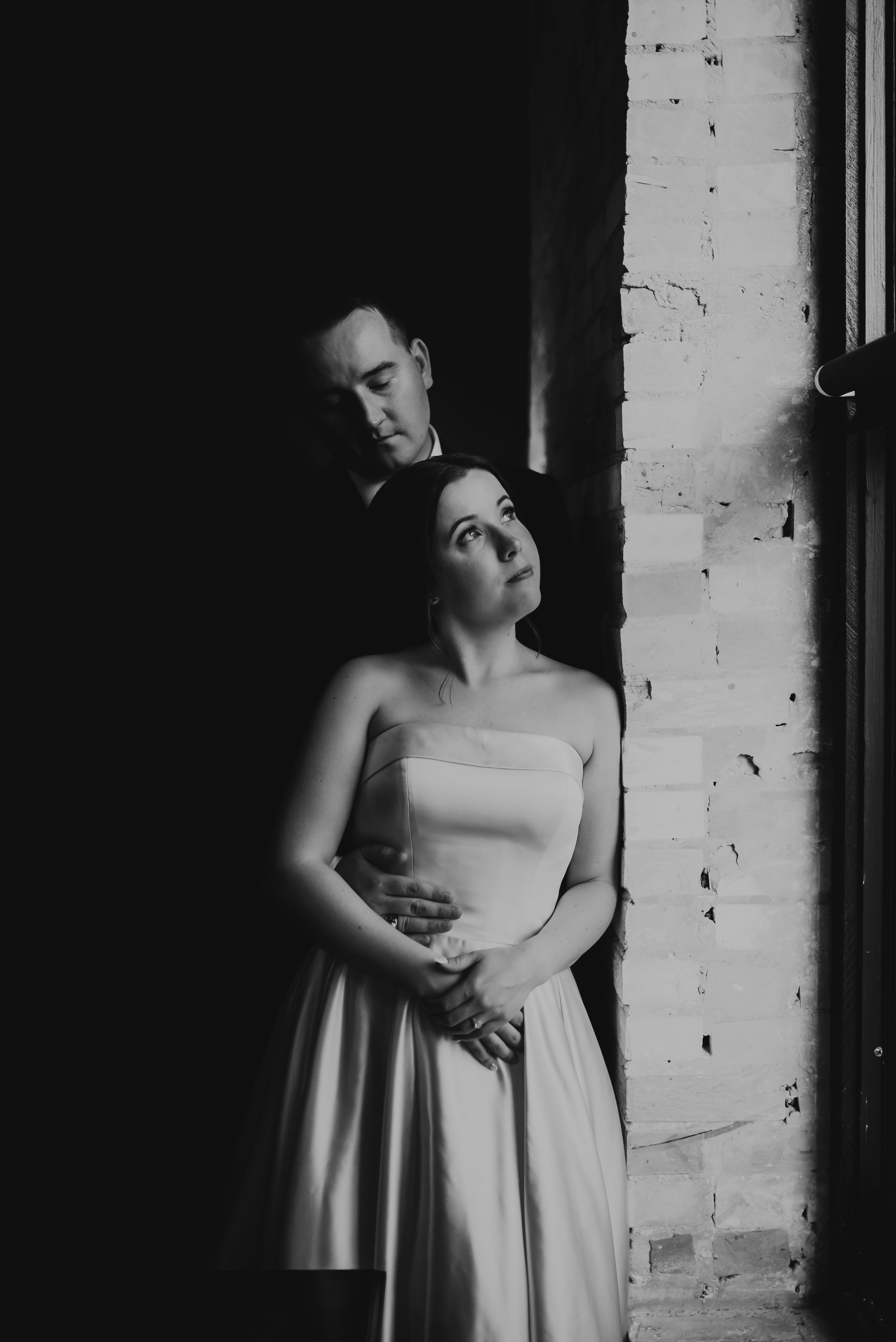 Black and white wedding portrait of bride and groom standing in dramatic light by brick wall.