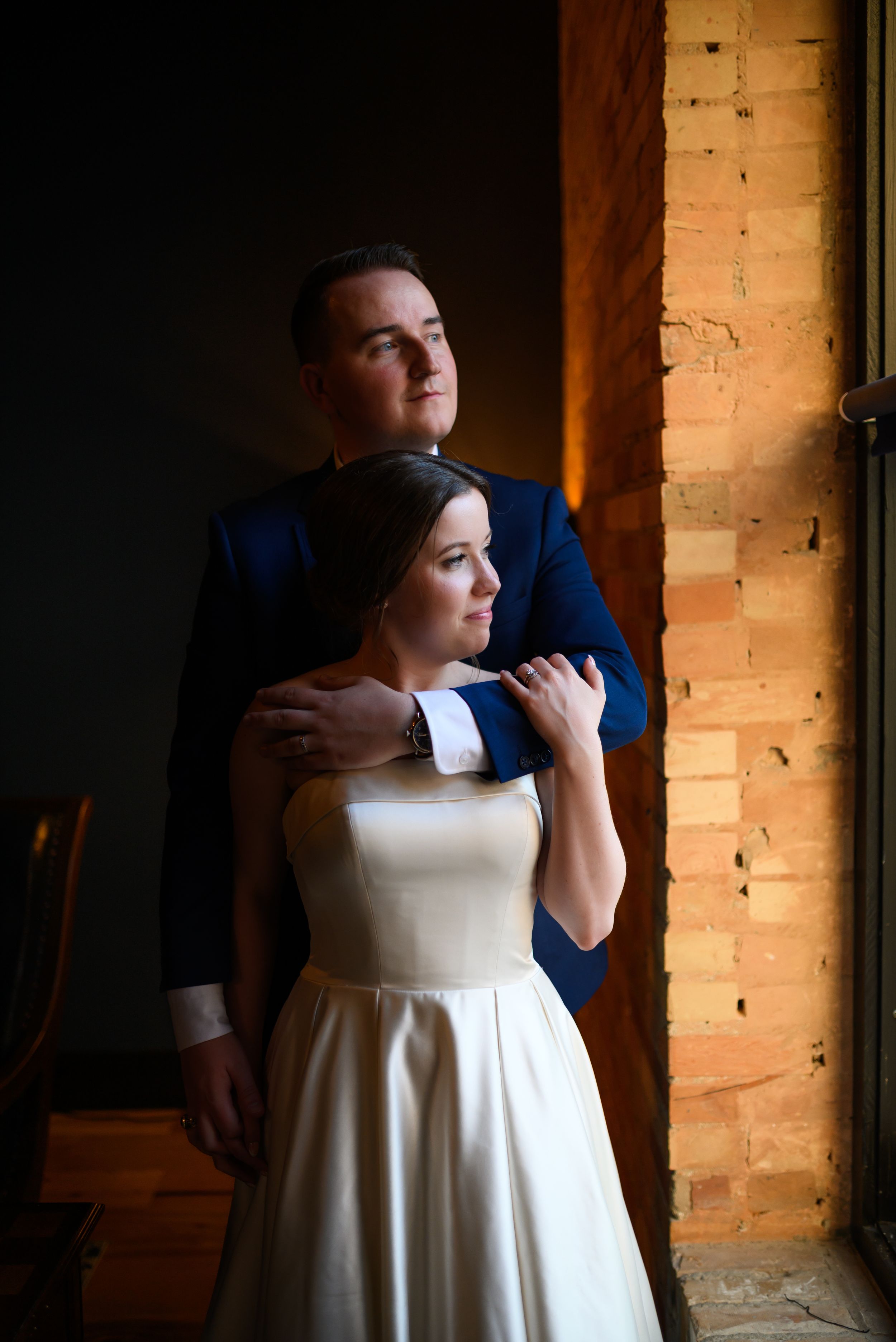 A bride in a white gown and groom in a blue suit embrace near a brick window in a romantic wedding portrait.