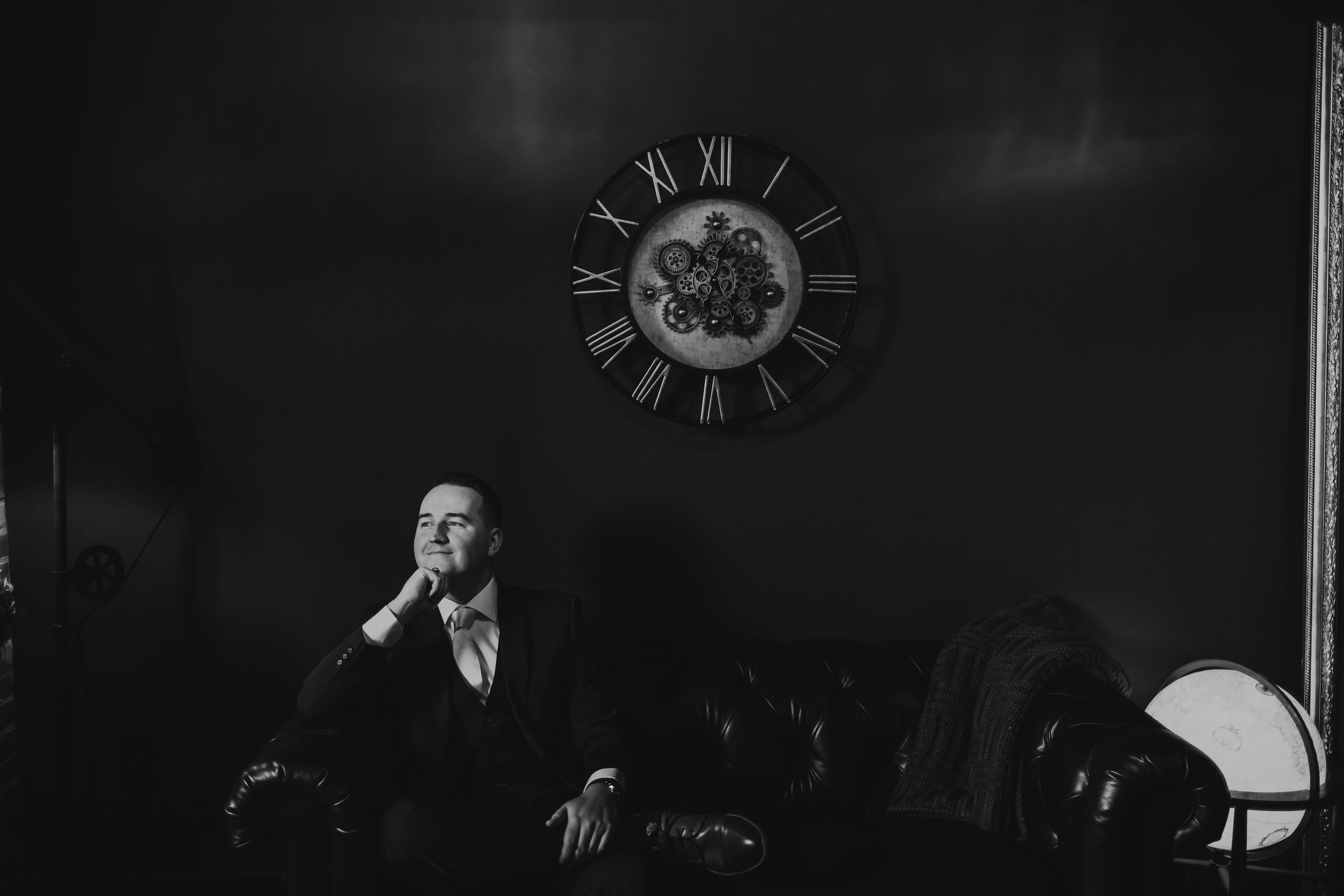 Black and white portrait of a person sitting on a leather sofa beneath a decorative wall clock in a dark room.