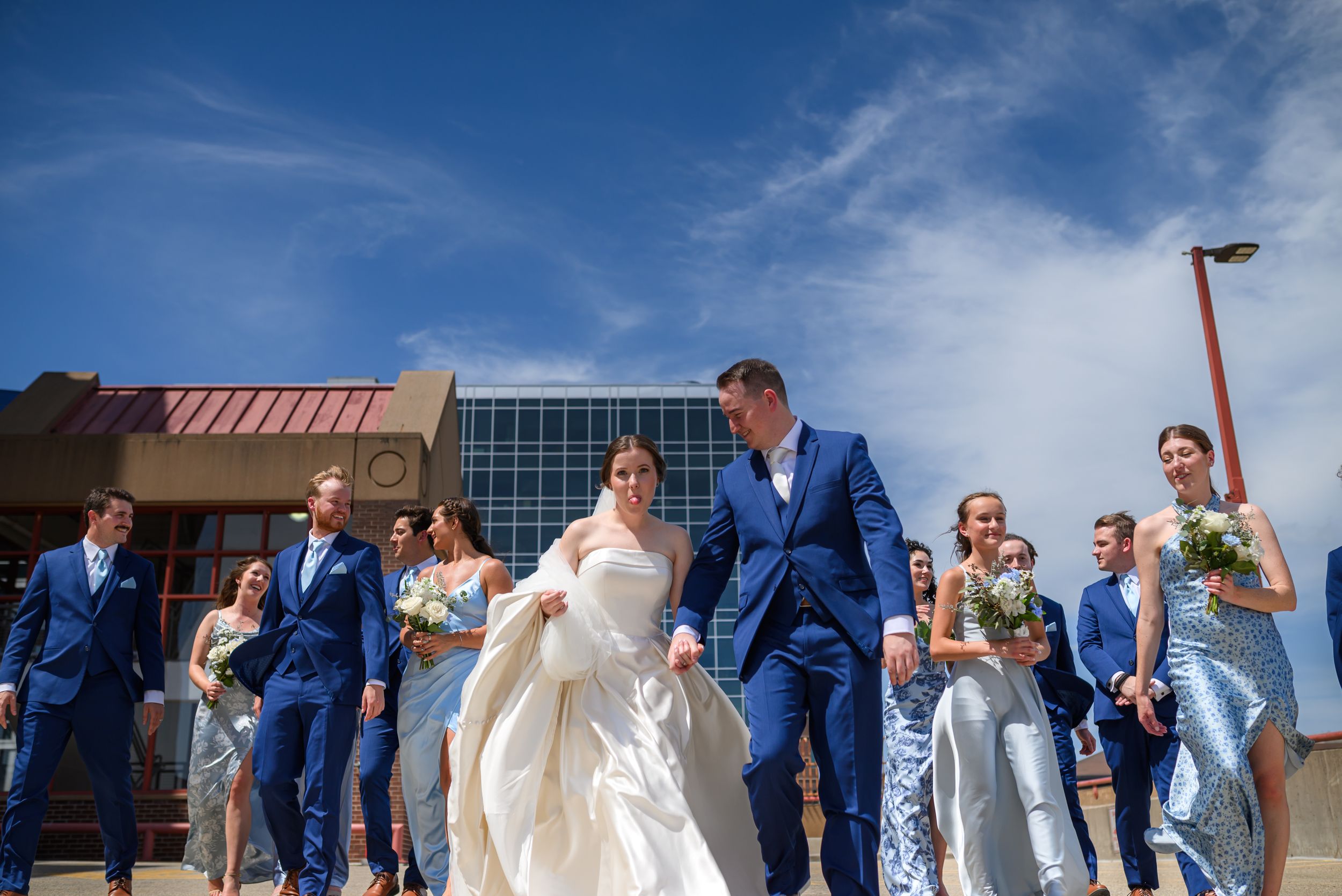 A wedding party in blue suits and silver dresses walks confidently outdoors under a bright blue sky.