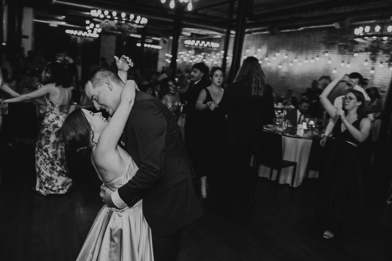Black and white photo of couple sharing romantic first dance at elegant wedding reception with guests celebrating in background.