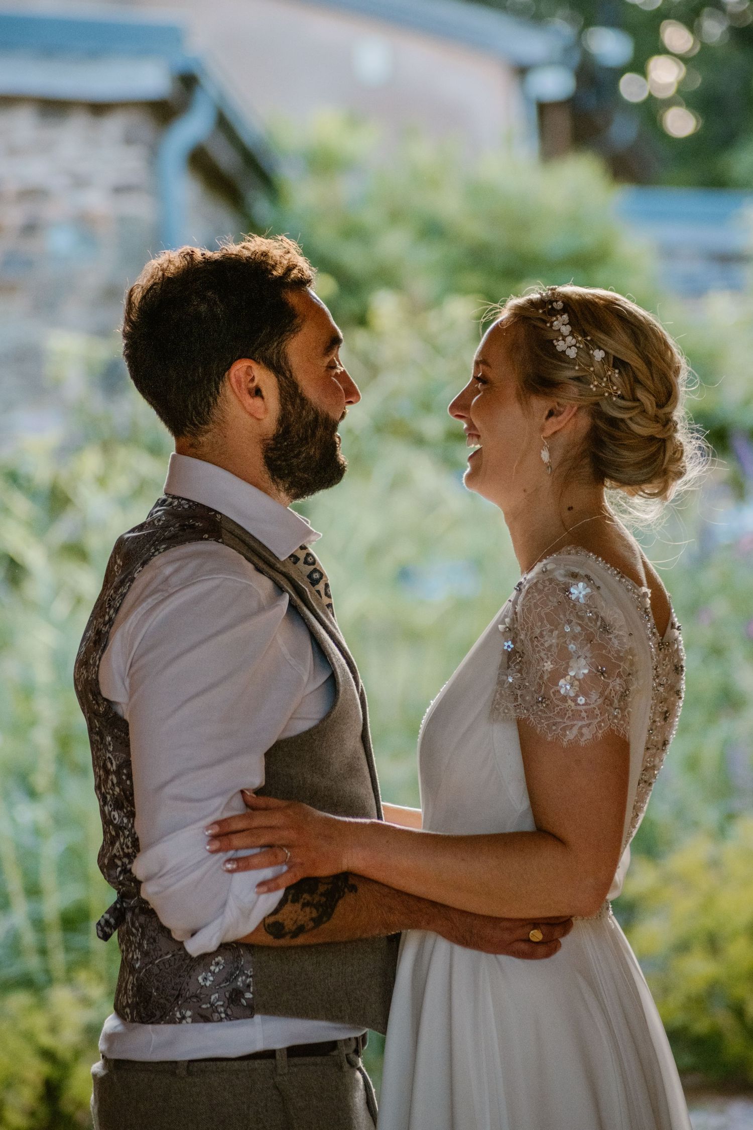 Bride and groom pose with two French Bulldogs under a stunning purple wisteria arch at their outdoor wedding.