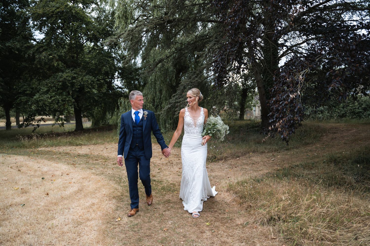 Bride in white gown and groom in blue suit holding hands walking along a rustic countryside path at Oxleaze Barn Wedding Venue.