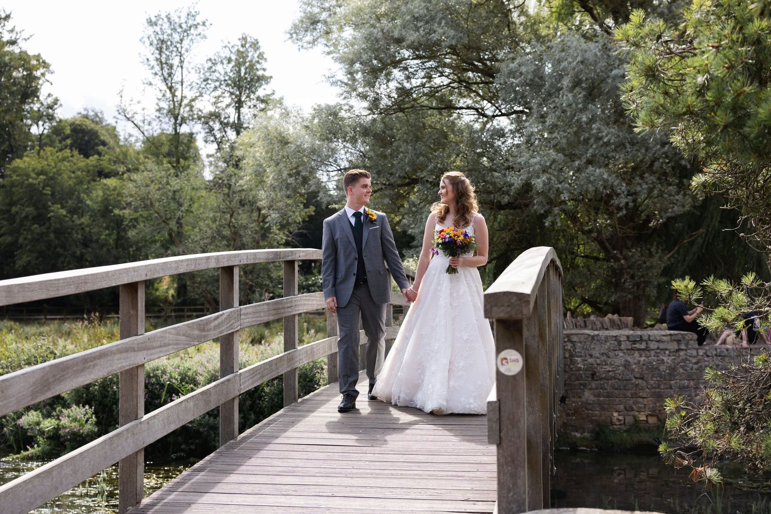 Bride and groom walk hand in hand across Bibury's wooden bridge surrounded by lush greenery.